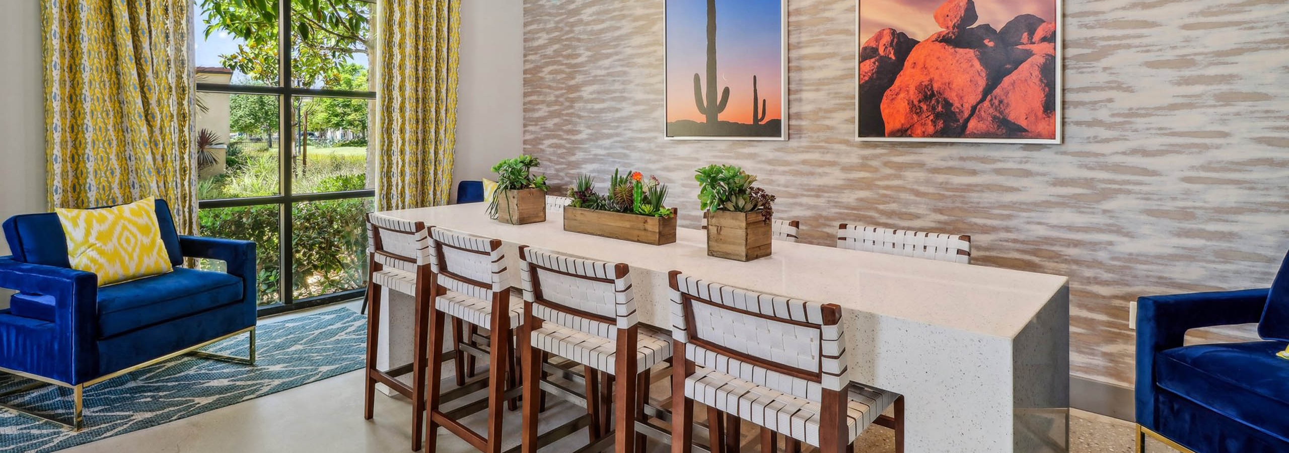 Clubroom at AMLI Camarillo apartments featuring a quartz-topped table, wood chairs, desert artwork and arched window with yellow drapes