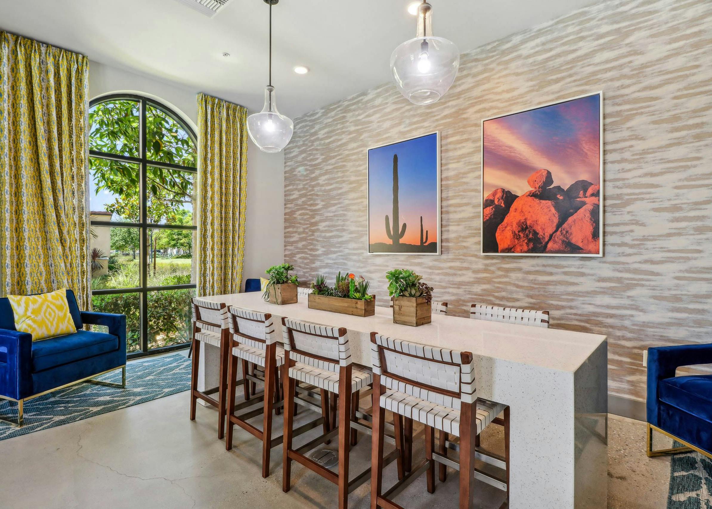 Clubroom at AMLI Camarillo apartments featuring a quartz-topped table, wood chairs, desert artwork and arched window with yellow drapes