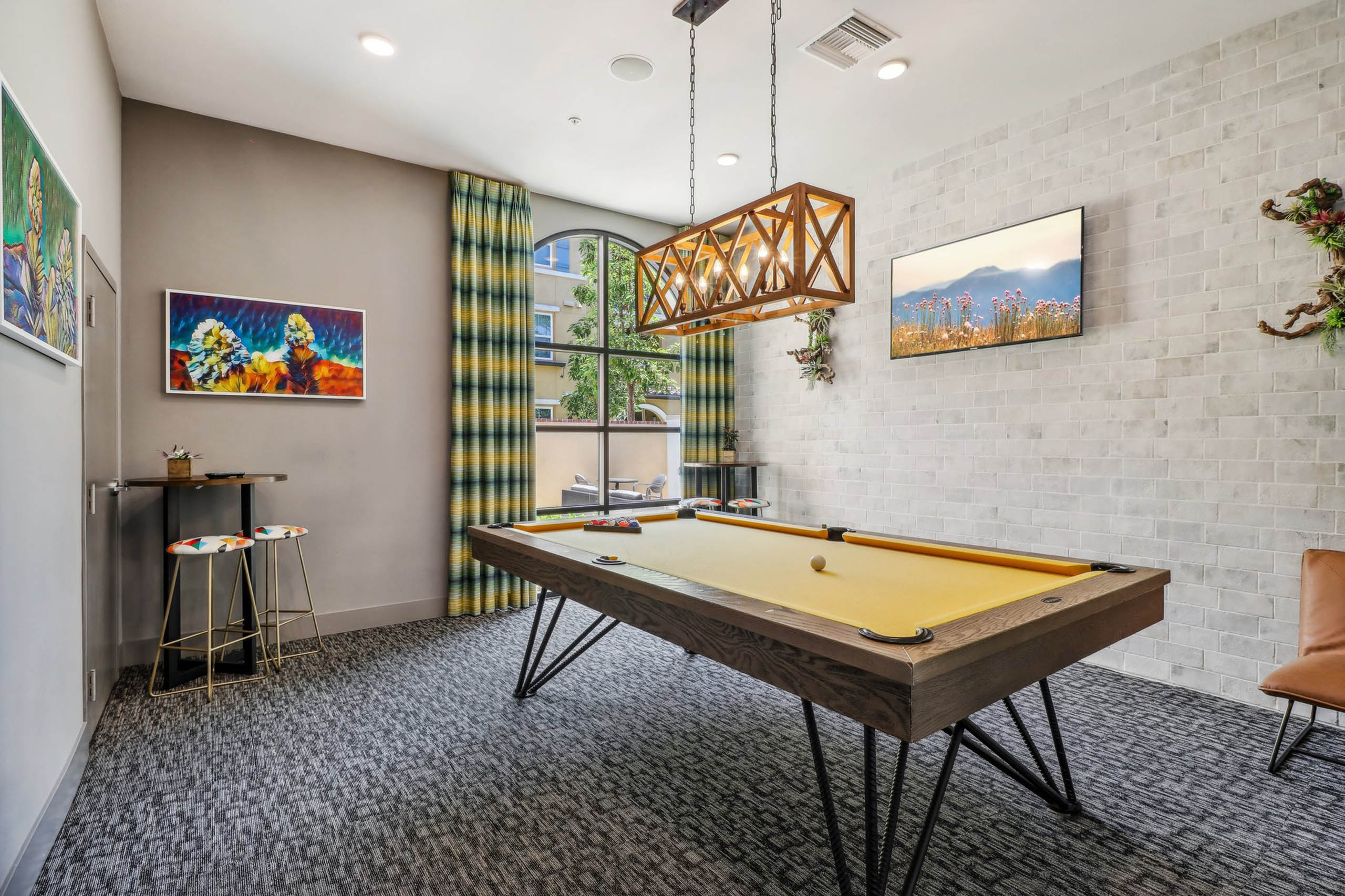 Game room at AMLI Camarillo apartments with yellow felt pool table, plaid curtains, colorful artwork and pendant chandelier over the table