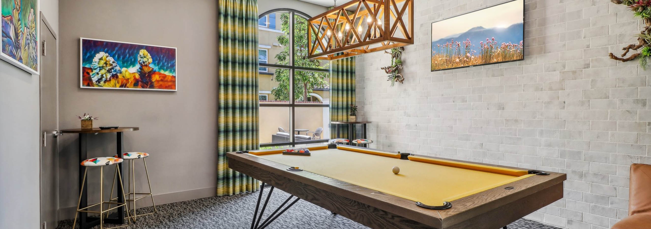 Game room at AMLI Camarillo apartments with yellow felt pool table, plaid curtains, colorful artwork and pendant chandelier over the table