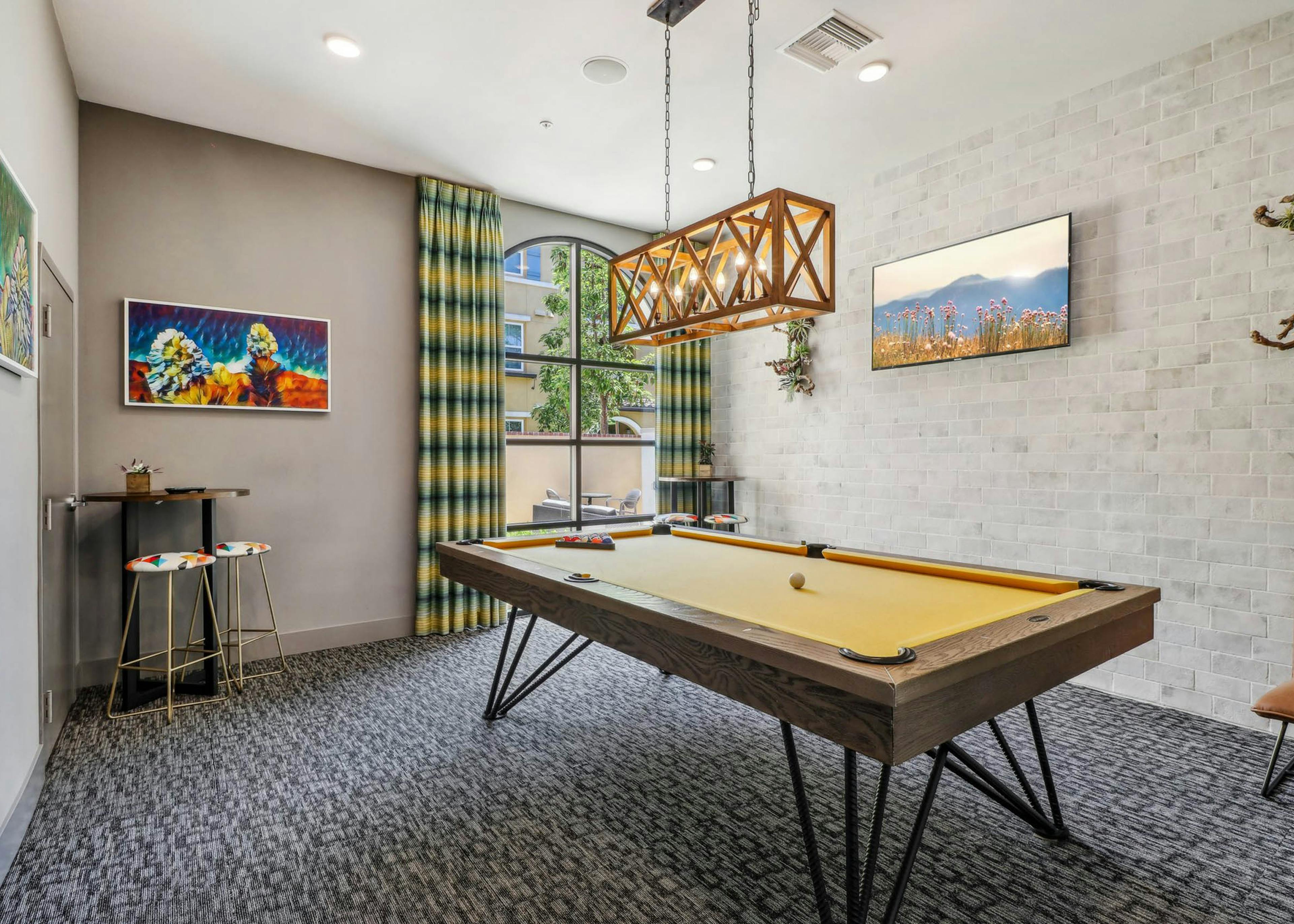 Game room at AMLI Camarillo apartments with yellow felt pool table, plaid curtains, colorful artwork and pendant chandelier over the table