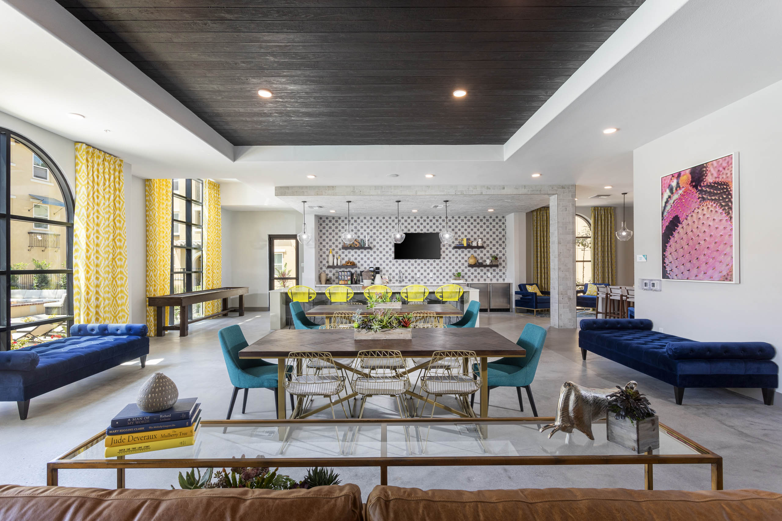 Wide view of AMLI Camarillo clubhouse with dining tables, lounge seating, shuffle board and patterned yellow curtains in a bright setting