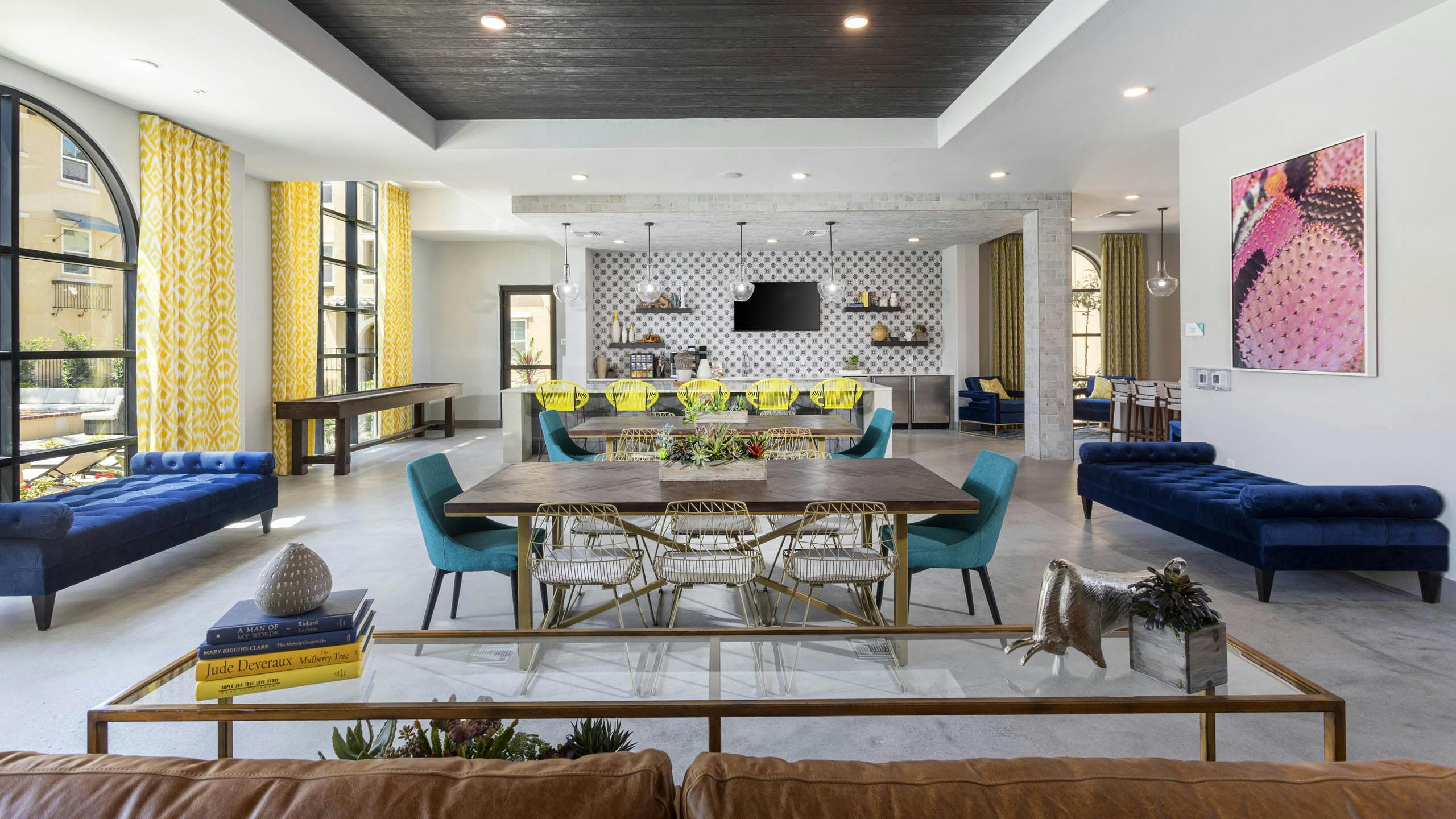 Wide view of AMLI Camarillo clubhouse with dining tables, lounge seating, shuffle board and patterned yellow curtains in a bright setting