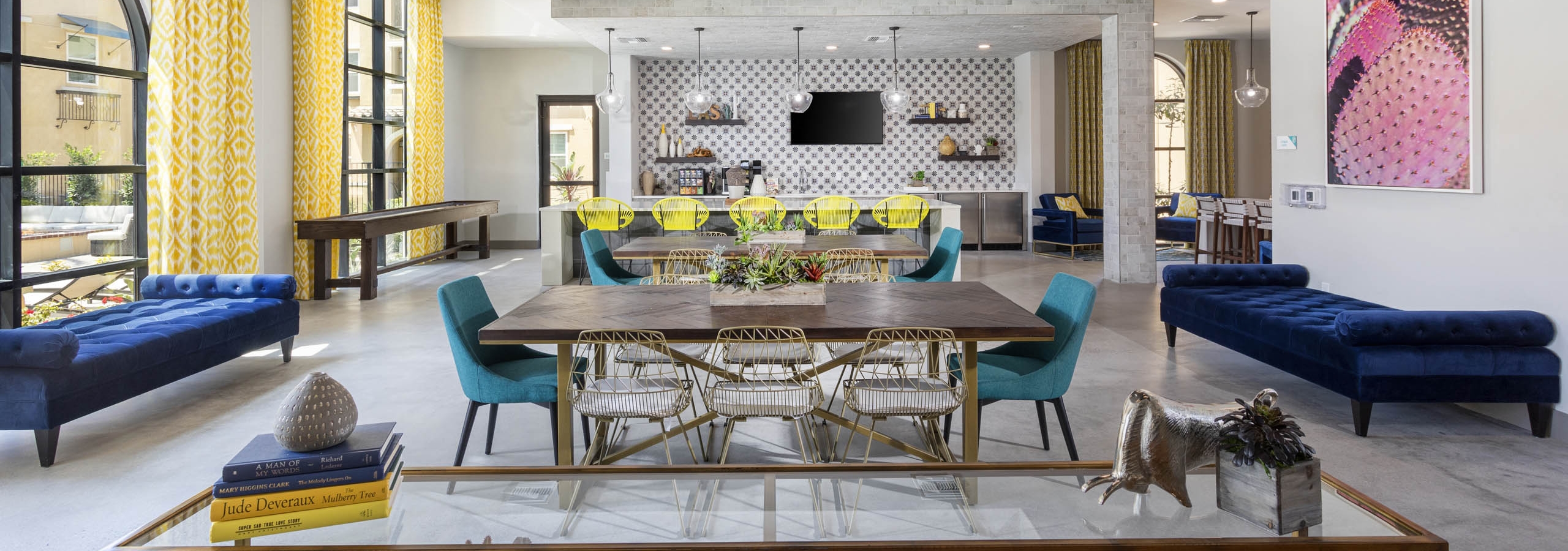 Wide view of AMLI Camarillo clubhouse with dining tables, lounge seating, shuffle board and patterned yellow curtains in a bright setting