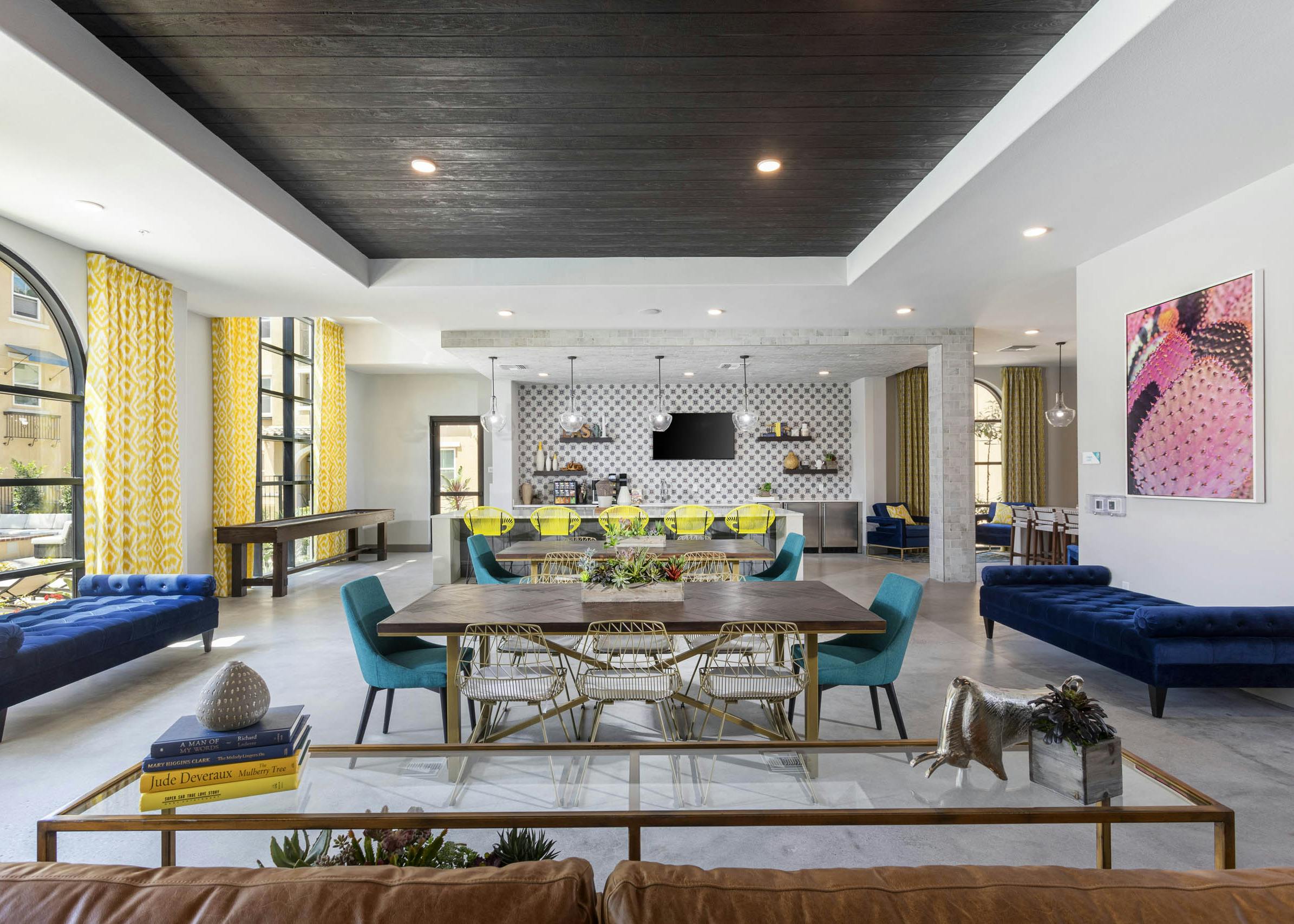Wide view of AMLI Camarillo clubhouse with dining tables, lounge seating, shuffle board and patterned yellow curtains in a bright setting