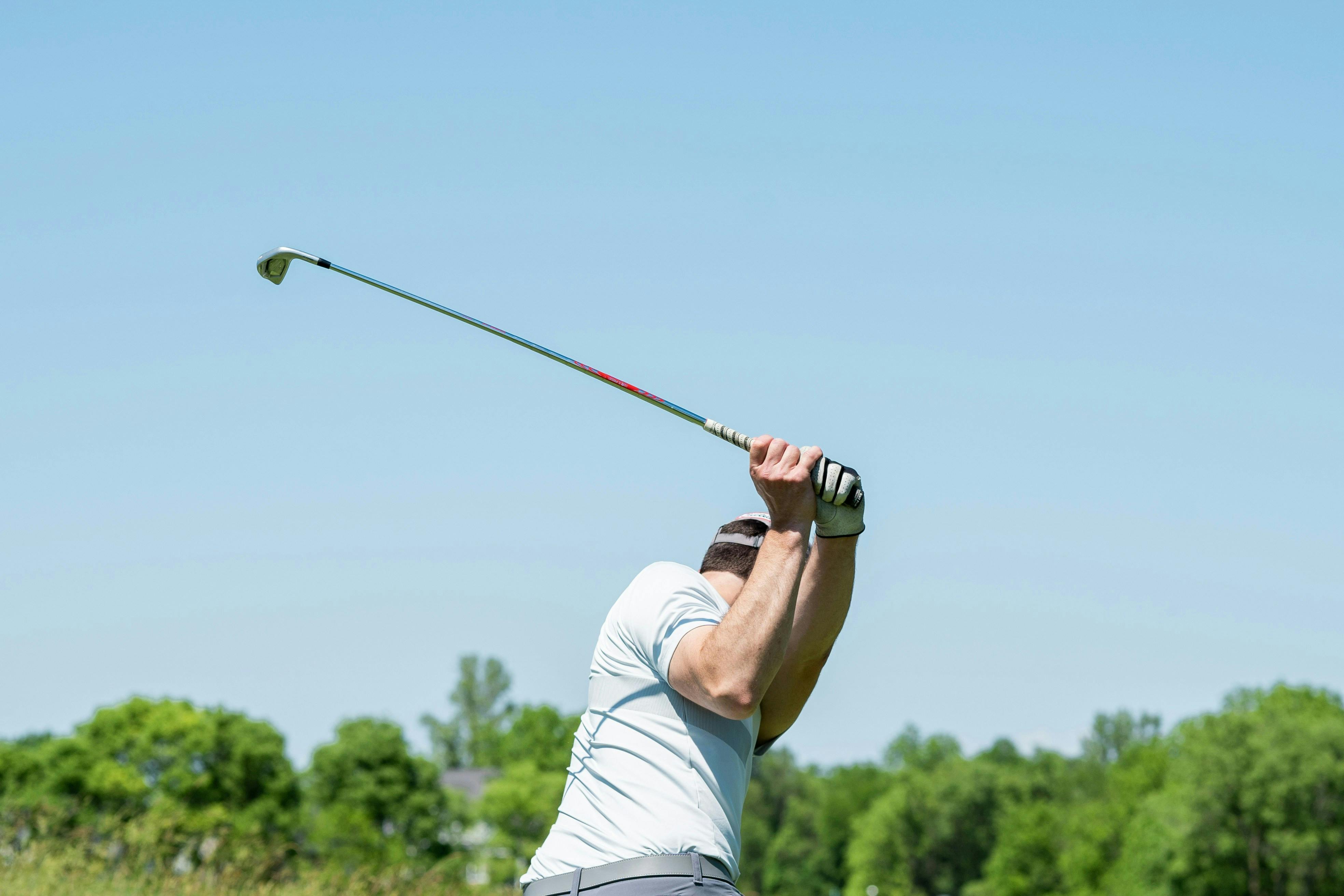 A close-up, low-angle shot of a male golfer's arms and shoulders at the top of his golf swing. He is wearing a white shirt and gloves, with the club held high against a clear, bright blue sky and a background of green trees.