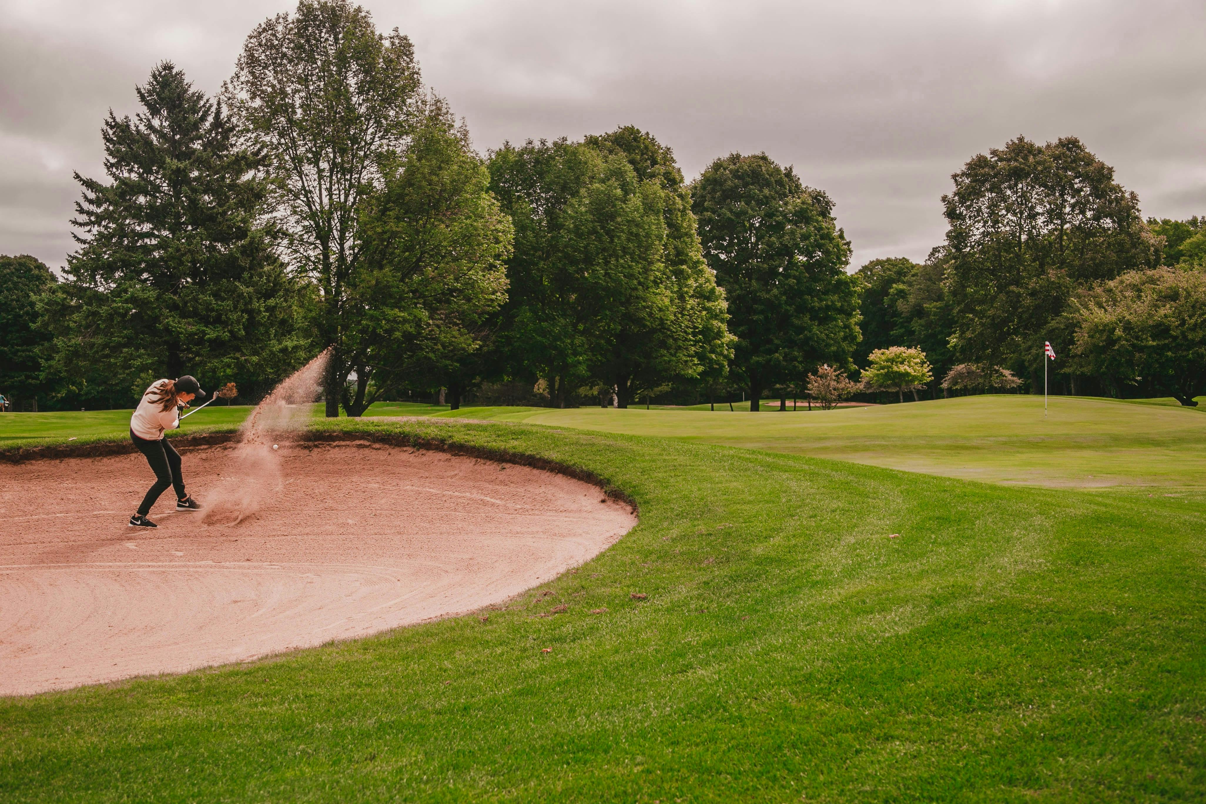 A female golfer is successfully hitting a shot out of a large sand trap or bunker, causing a visible spray of sand into the air. The bunker curves alongside a bright green fairway, with a line of tall, dark trees in the background under an overcast sky.