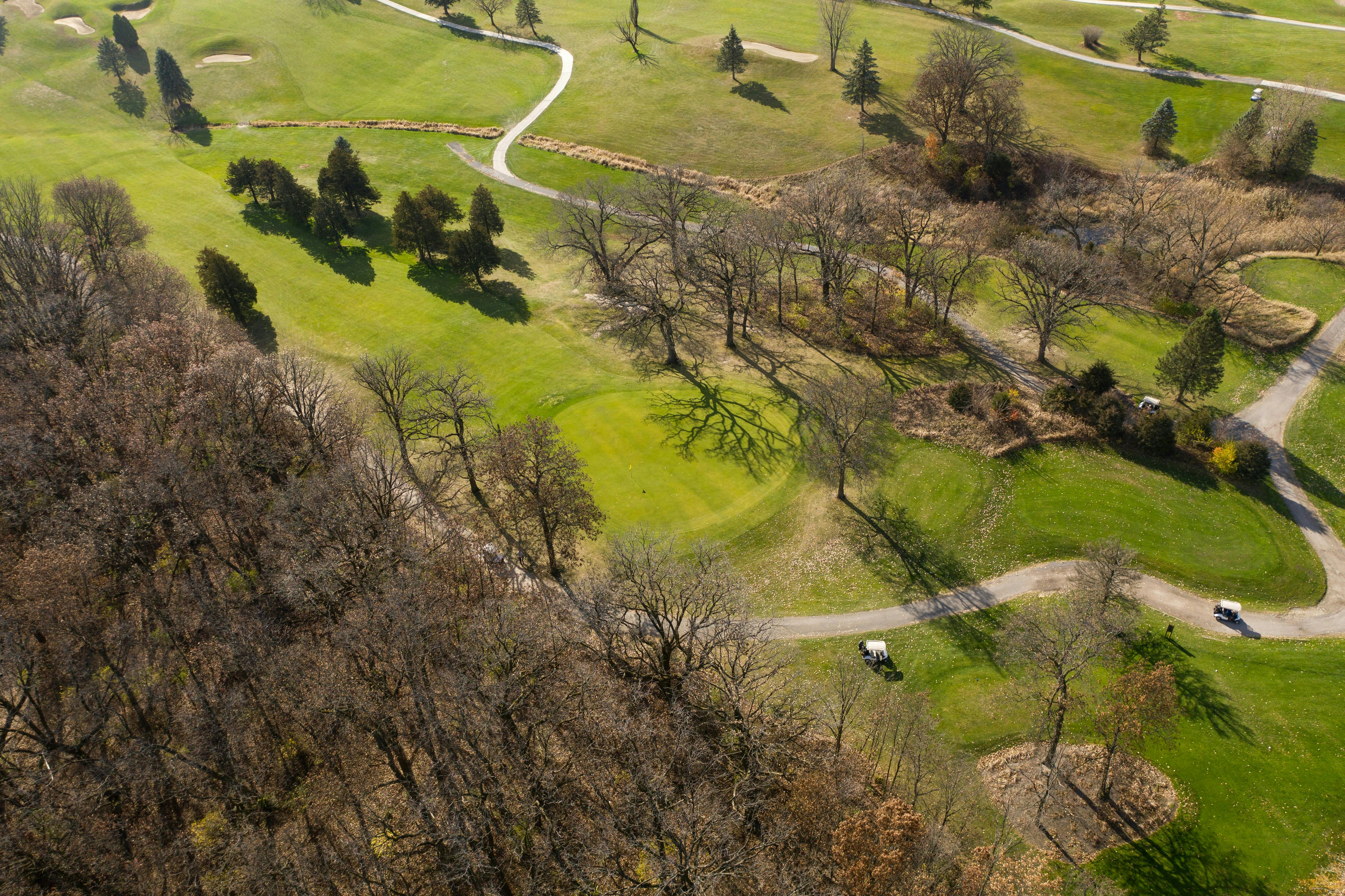 An aerial view of a golf course during the late fall or early winter. The grass is still green, but many of the trees are bare and leafless, casting long shadows. A winding white cart path crosses the fairways, and a couple of golf carts are visible.