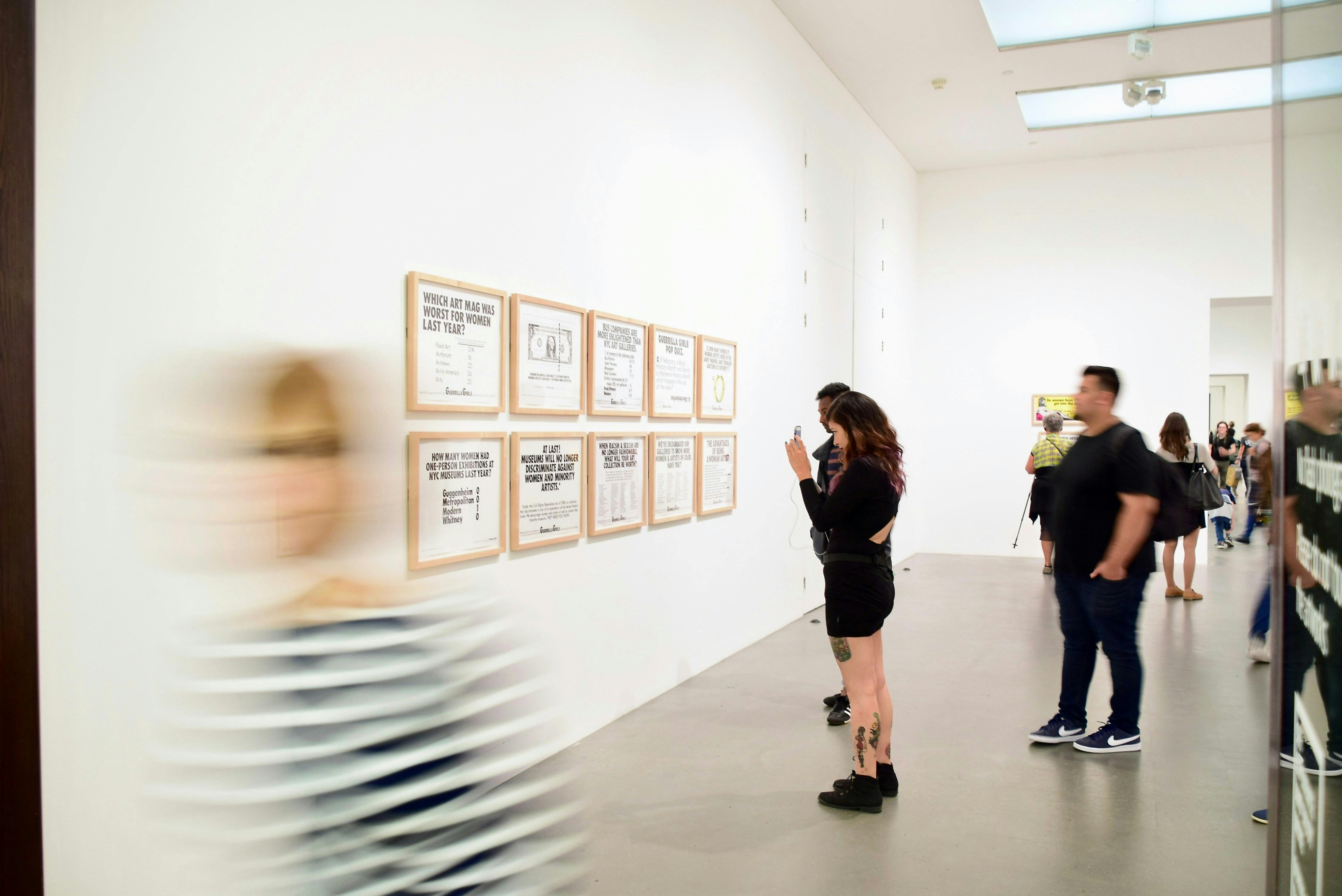A modern, white-walled art gallery scene. A row of framed text-based art pieces is displayed on the wall. A woman in a black dress is standing in the foreground, taking a photo of the art with her phone. Several other patrons are visible in the background, while a person in a striped shirt in the far left foreground is blurred by motion.

