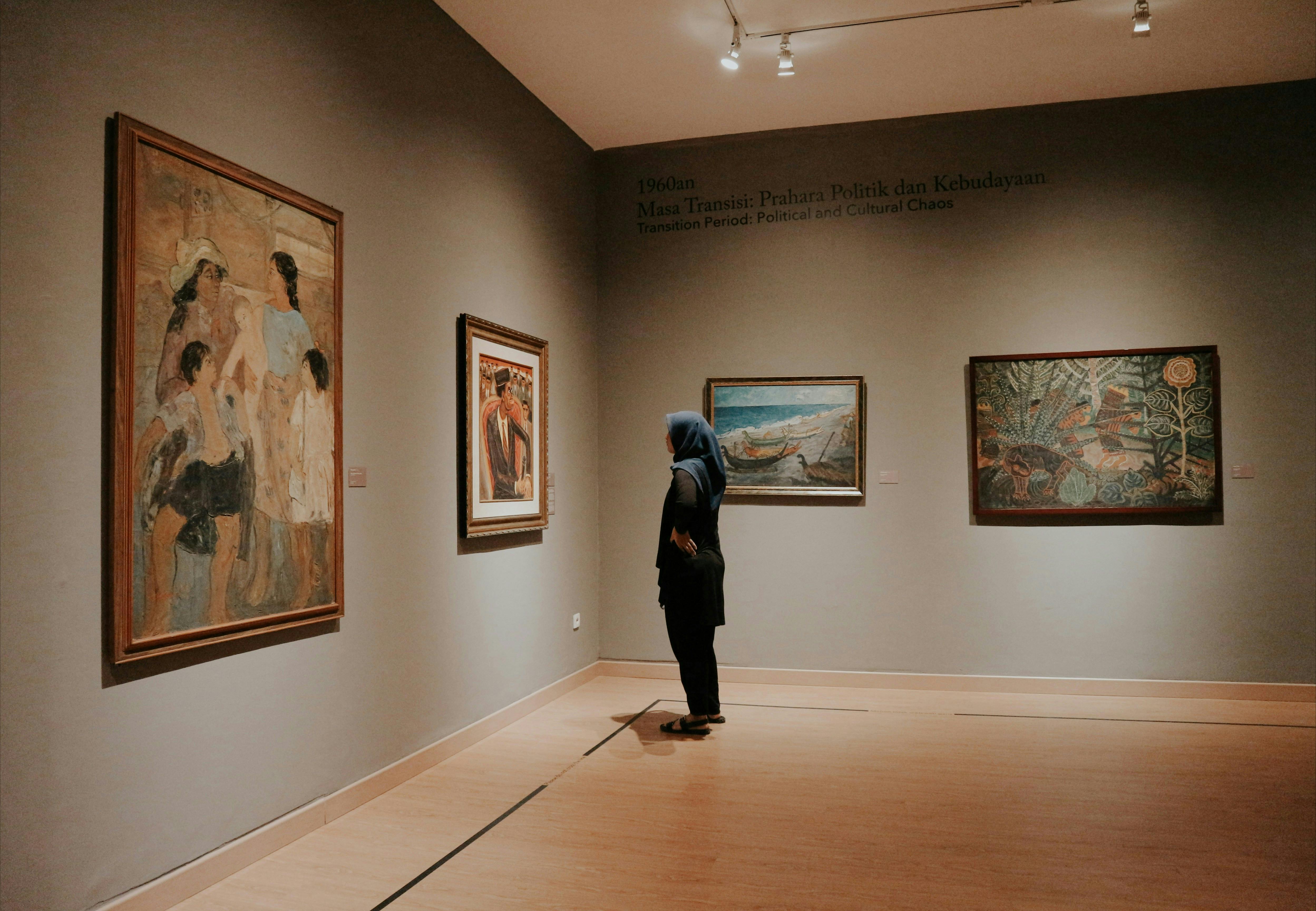 A person wearing a dark blue hijab and all-black clothing stands alone in a dimly lit art gallery, observing a painting on a muted brown wall. The wall also features three other paintings, with text near the ceiling in a foreign language (likely Indonesian) and English describing a "Transition Period: Political and Cultural Chaos."