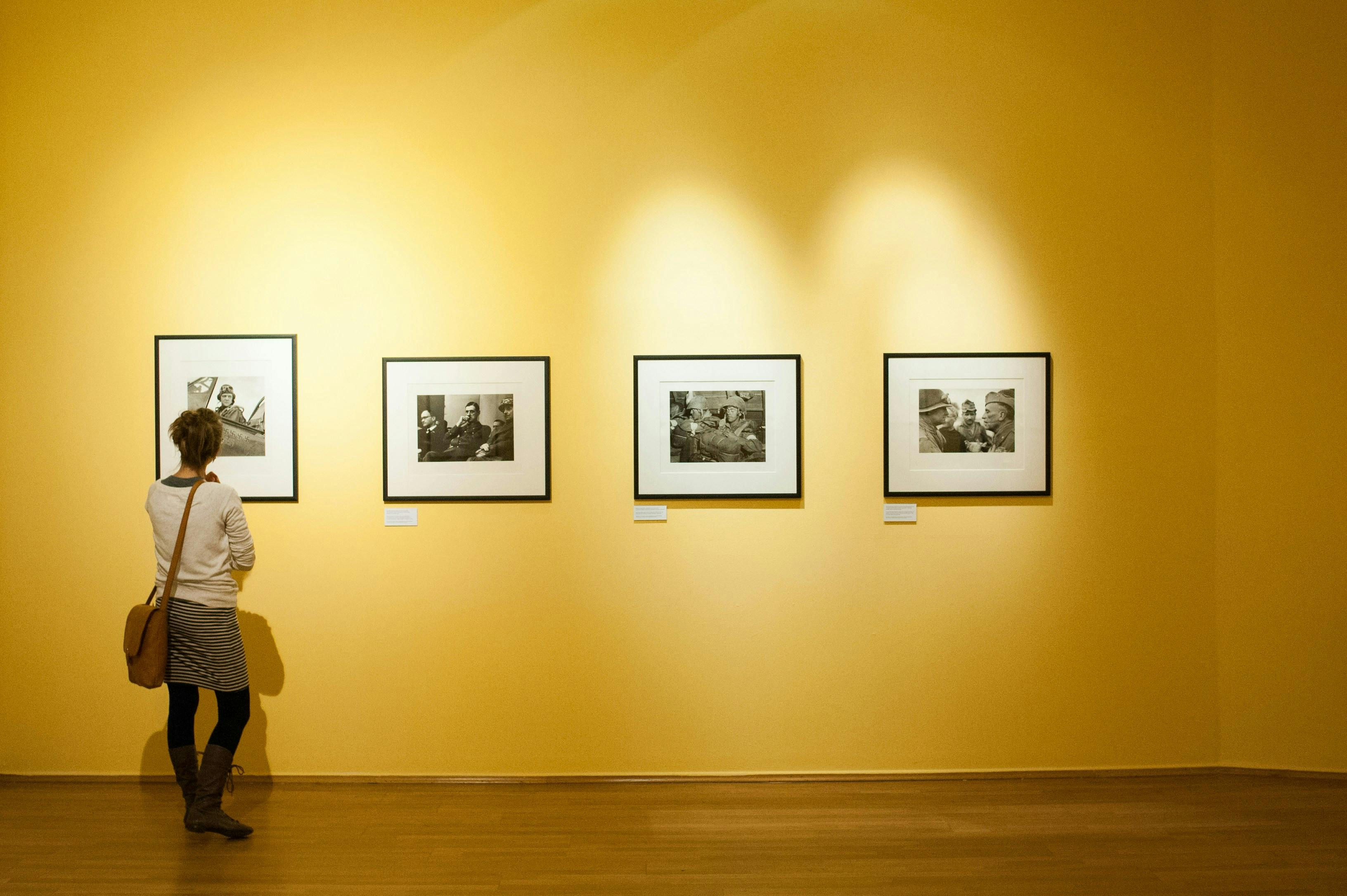 A woman viewed from behind, wearing a striped skirt and a white sweater, stands on a wooden floor, attentively looking at one of four framed, black-and-white photographs displayed in a row on a large, brightly lit mustard-yellow wall.