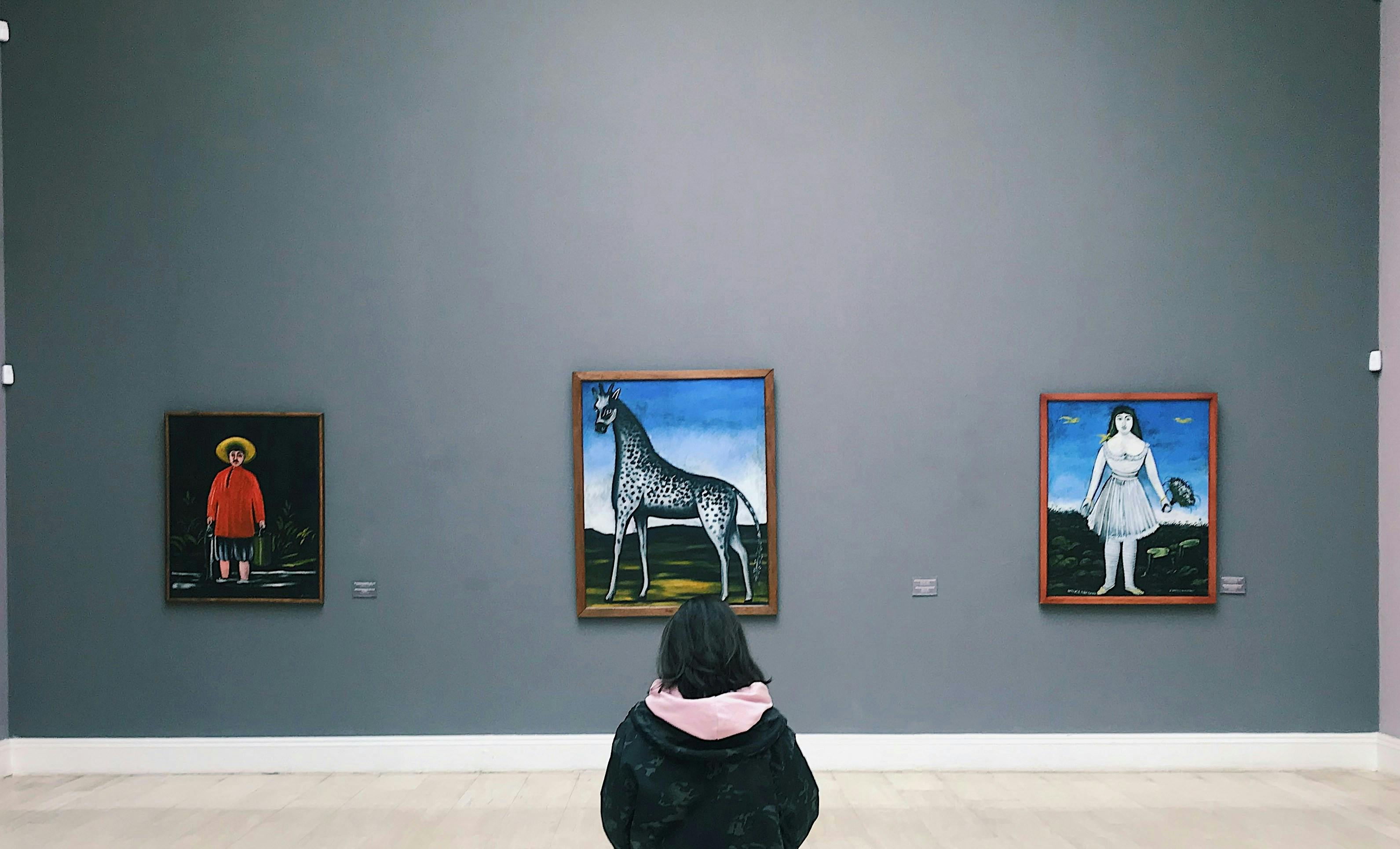 A person with dark hair in a black and pink hooded jacket stands in an art gallery, viewing three framed paintings mounted on a wide, featureless grey wall. The paintings appear to be in a surrealist or folk-art style, depicting a child in a red jacket, a spotted giraffe, and a figure in a white dress.