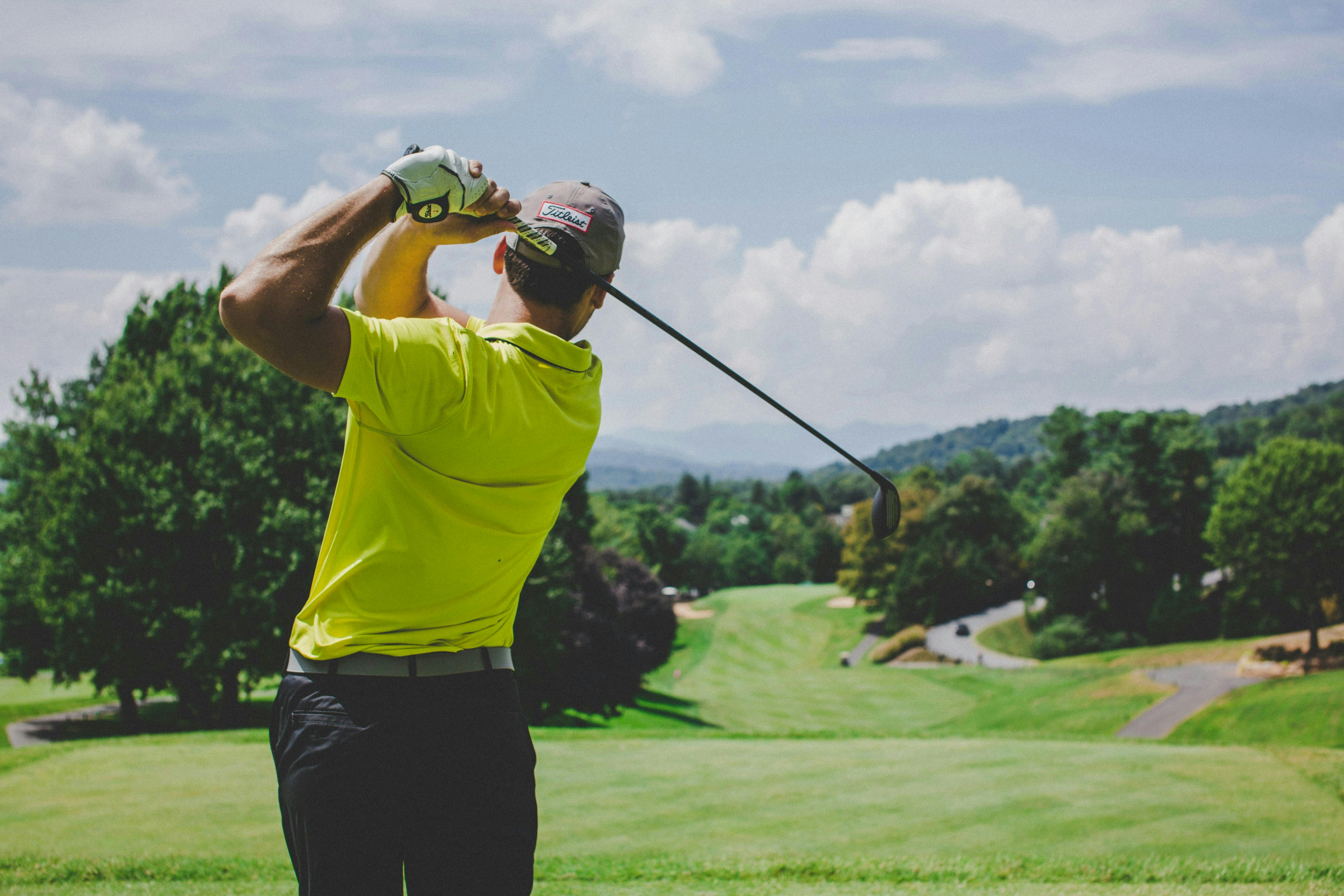A male golfer, viewed from behind, wearing a bright yellow-green polo shirt and a cap, is at the top of his backswing with a driver. The image overlooks a sloping, lush green golf course framed by trees, with rolling hills and mountains in the far distance under a blue and white cloudy sky.

