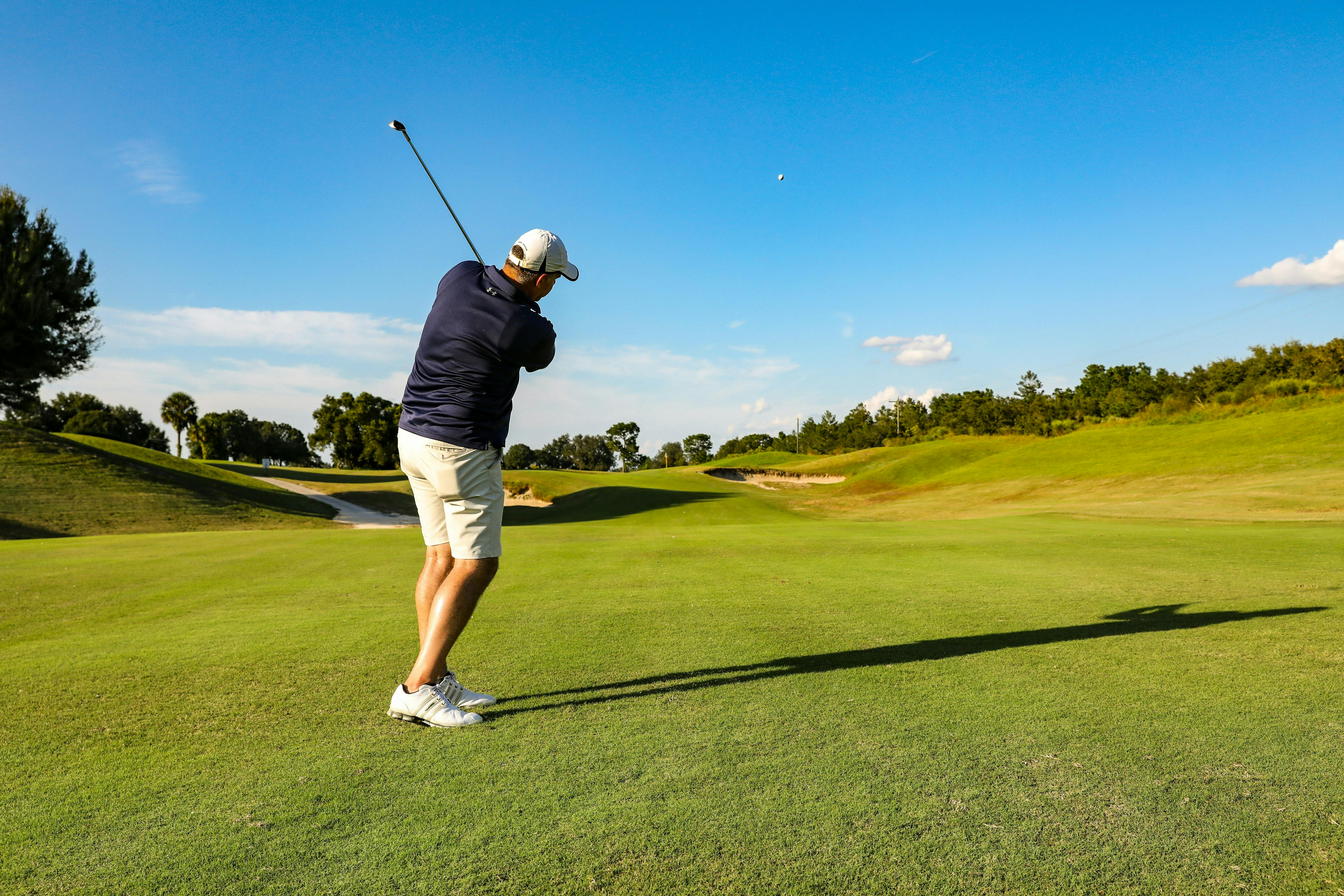 A male golfer in a navy shirt, light shorts, and a white hat is captured mid-swing on a sunny, wide-open fairway. His shadow stretches long in the afternoon sun, and a white golf ball is visible in the air against a deep blue sky with a few clouds.

