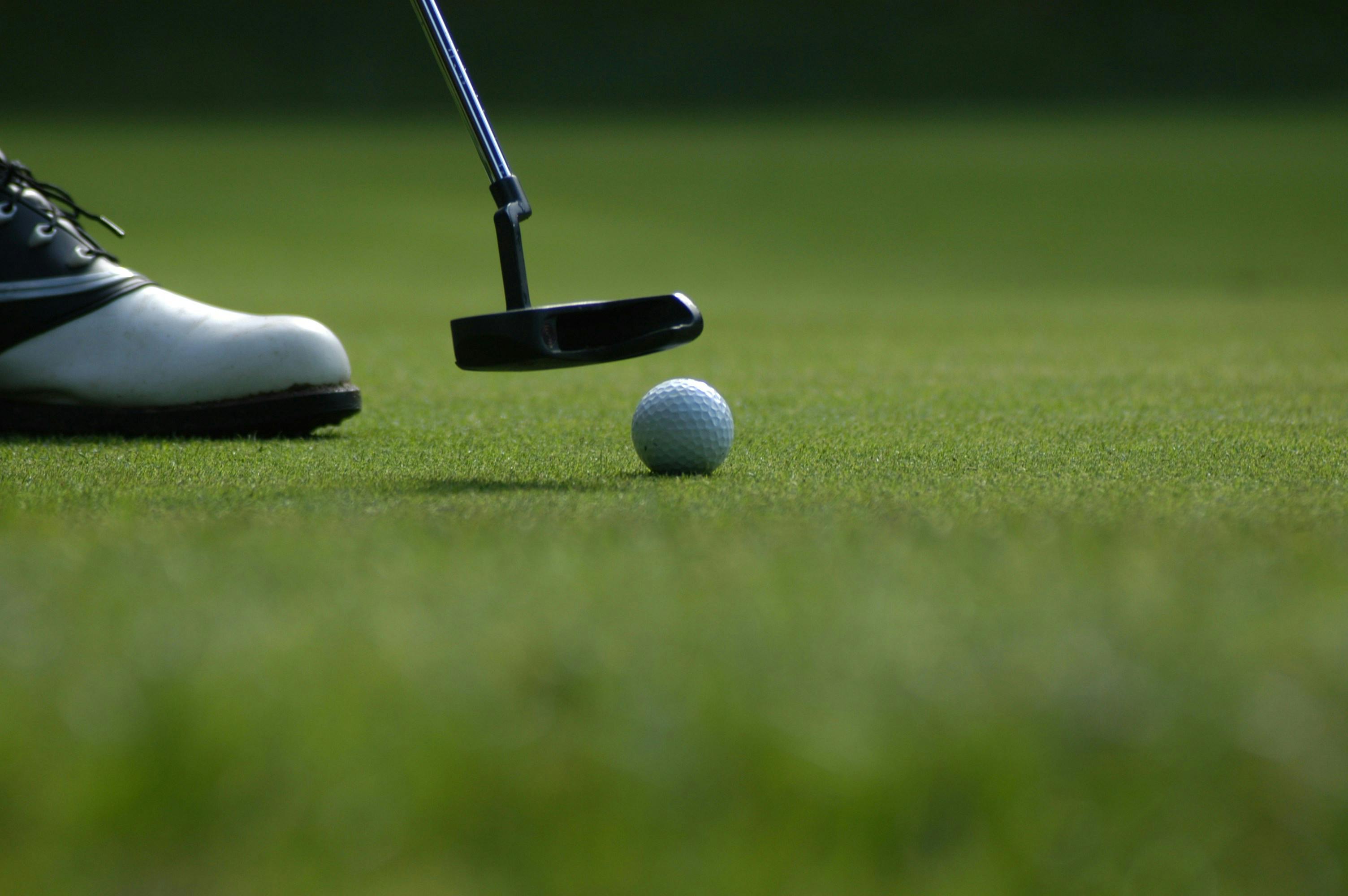 A low, close-up shot of a putter hovering just over a white golf ball on a finely manicured green. The toe of a white and black golf shoe is visible on the far left. The green is shaded, and the focus is sharp on the ball and club.
