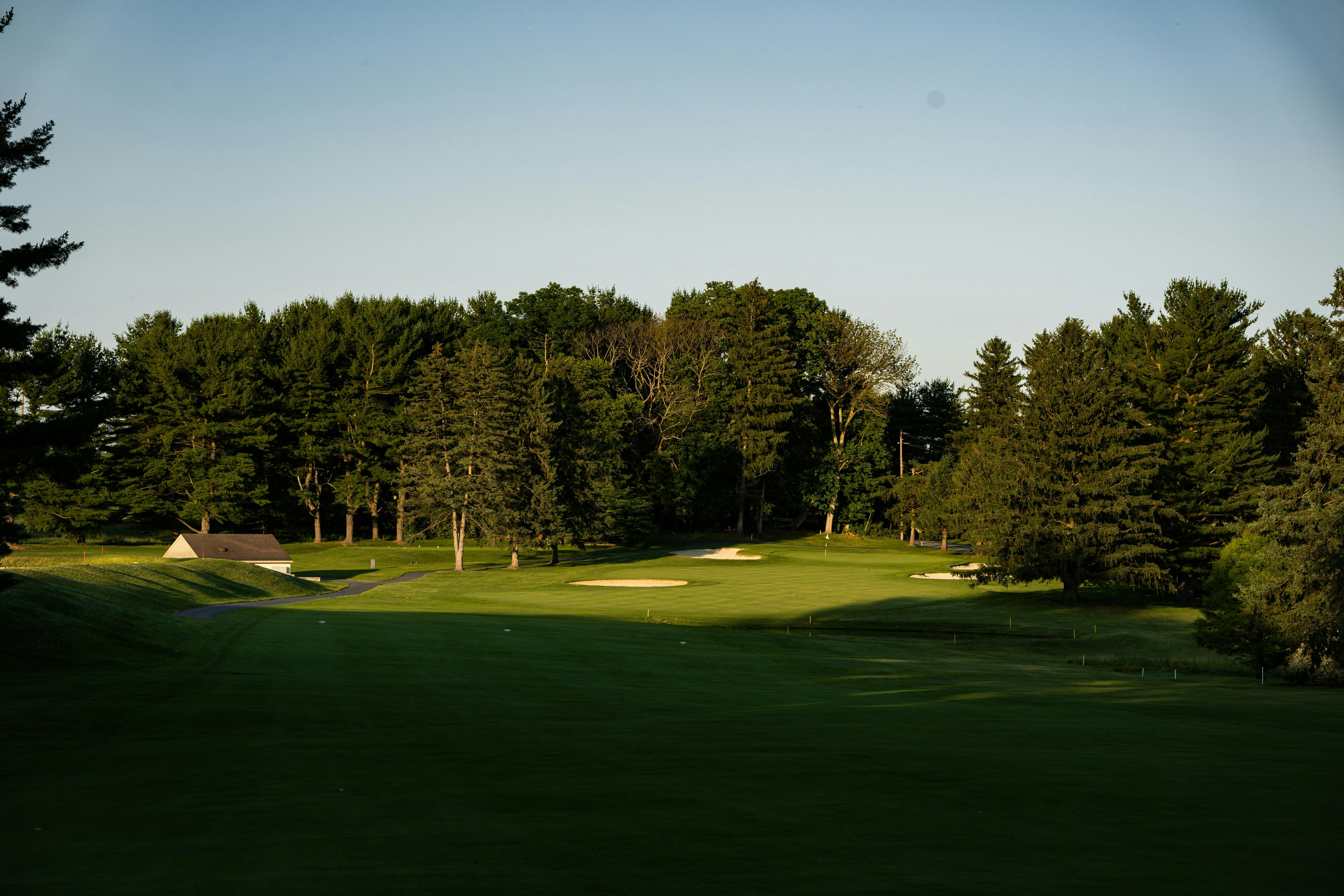 A wide-angle view of a golf course hole. The foreground is cast in deep shadow, while the middle ground, featuring a green with bunkers, is brightly lit. The scene is bordered by a dense line of various evergreen and deciduous trees, with a small utility building on the left.