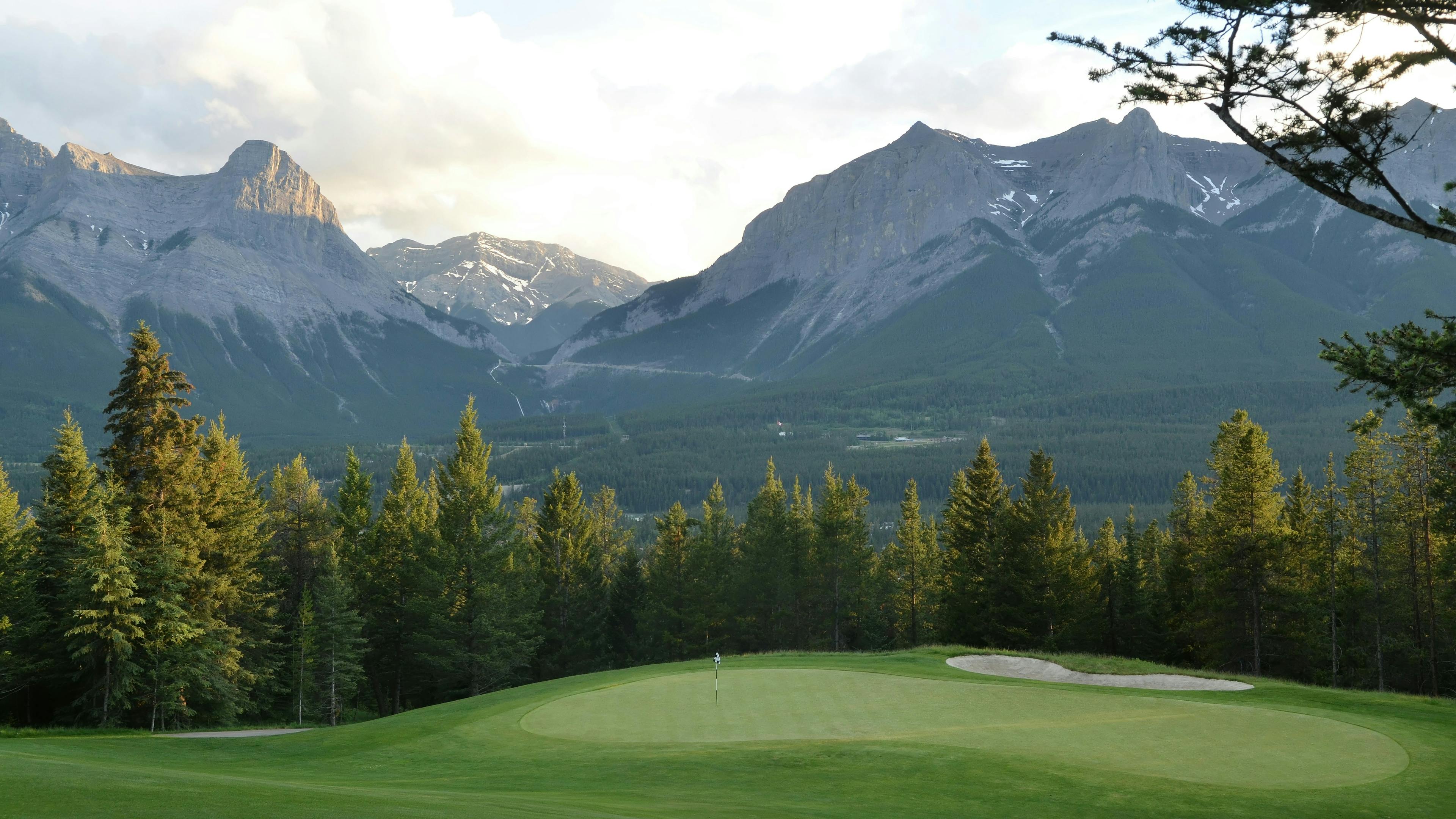 A scenic view of a golf green and bunker set against a dramatic backdrop of towering, rugged mountains with patches of snow. A dense line of evergreen trees separates the green from the mountains. The sky above is bright with sunset light and some clouds.