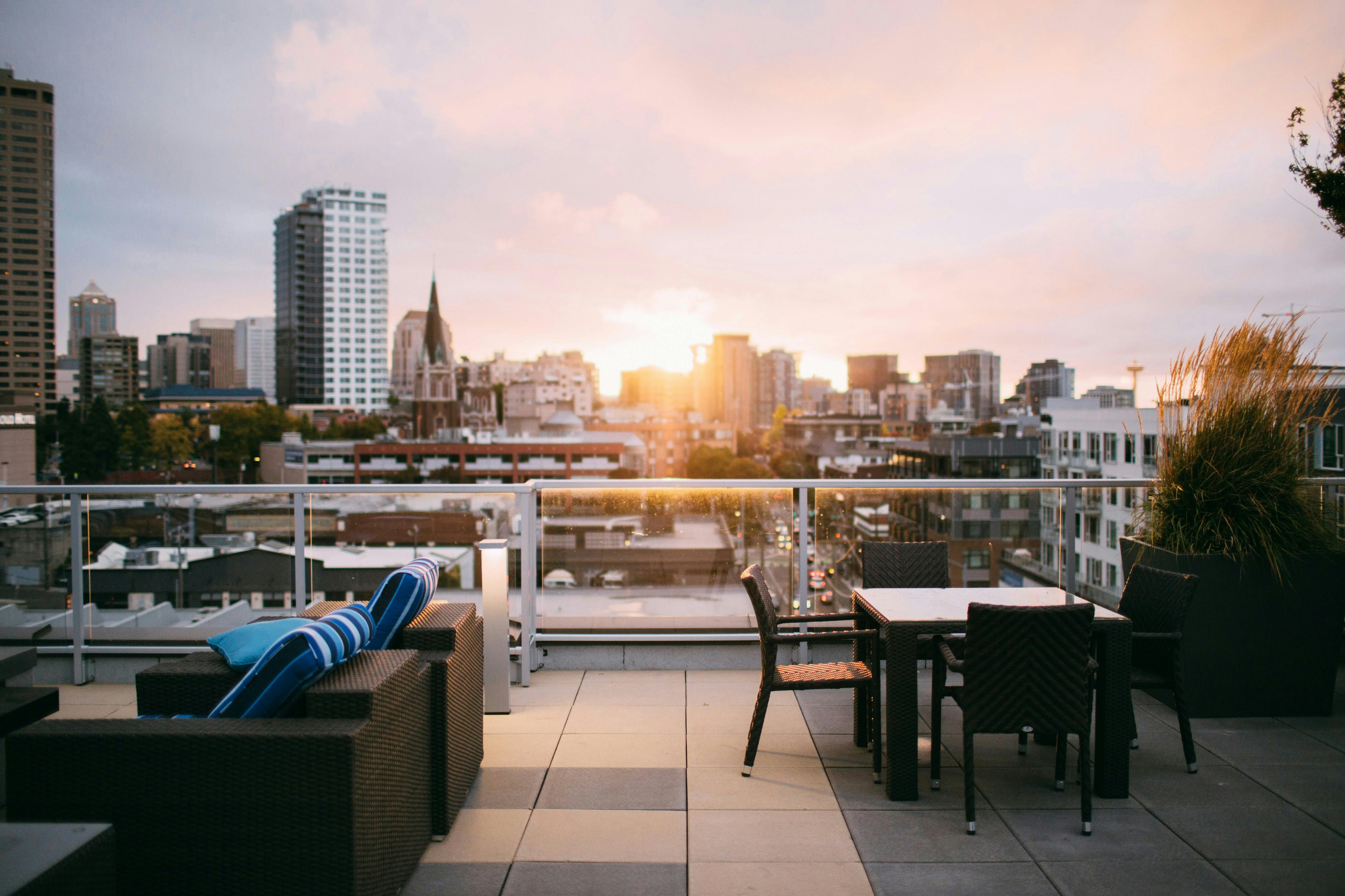 An inviting outdoor rooftop living area at sunset. The deck features dark woven patio furniture and a dining table. The sky is filled with soft, warm colors from the sunset over the blurred cityscape of Seattle, which includes a church spire and modern high-rises.