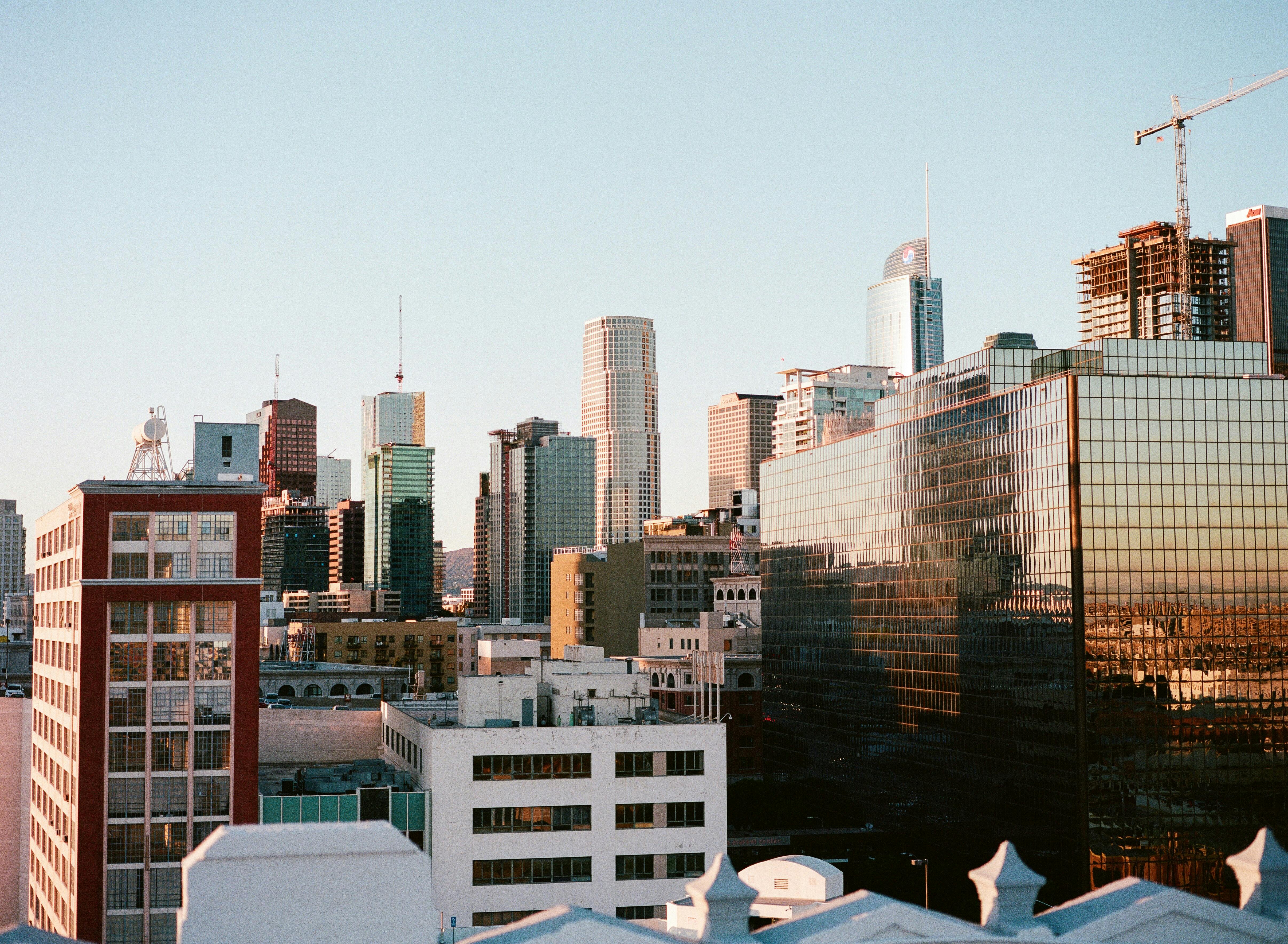 A close-up view of a dense cluster of downtown Seattle skyscrapers under a pale blue sky. The golden light of the setting sun warmly reflects off the glass facade of a large, angular building in the foreground on the right, contrasting with the dark red and grey colors of the surrounding structures.