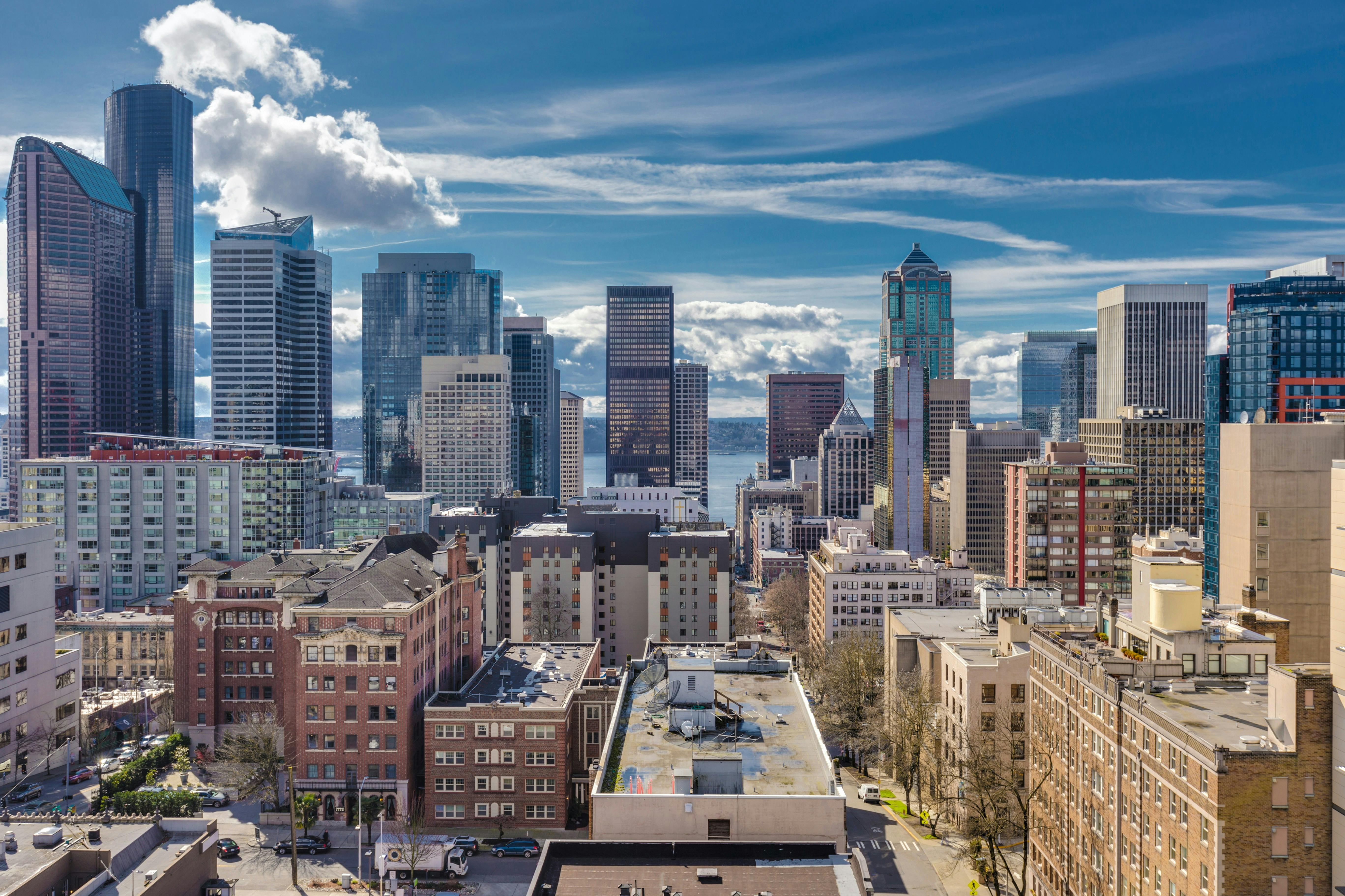 A wide view of the dense Seattle skyline on a bright, partially cloudy day. The image captures several layers of buildings, from the older, lower-rise residential and commercial buildings in the foreground to the cluster of modern, reflective skyscrapers in the background, with a glimpse of water (likely the Puget Sound) between them.
