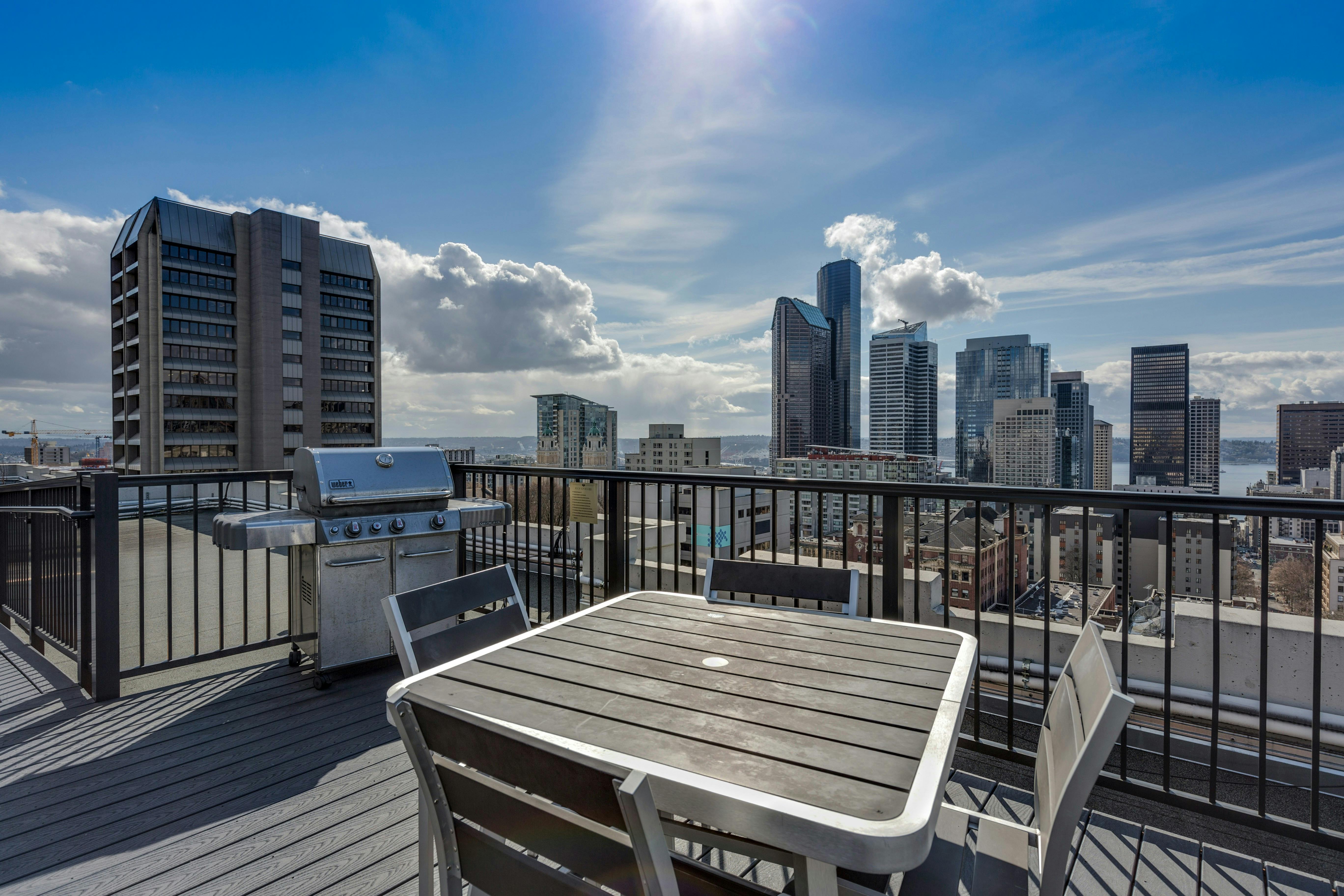 A brightly lit, sunny rooftop deck with an outdoor dining table and chairs in the foreground, and a large stainless steel grill on the left. A black railing encloses the space, offering a panoramic view of the downtown Seattle skyline, including the distinctive curved tops of some modern skyscrapers.