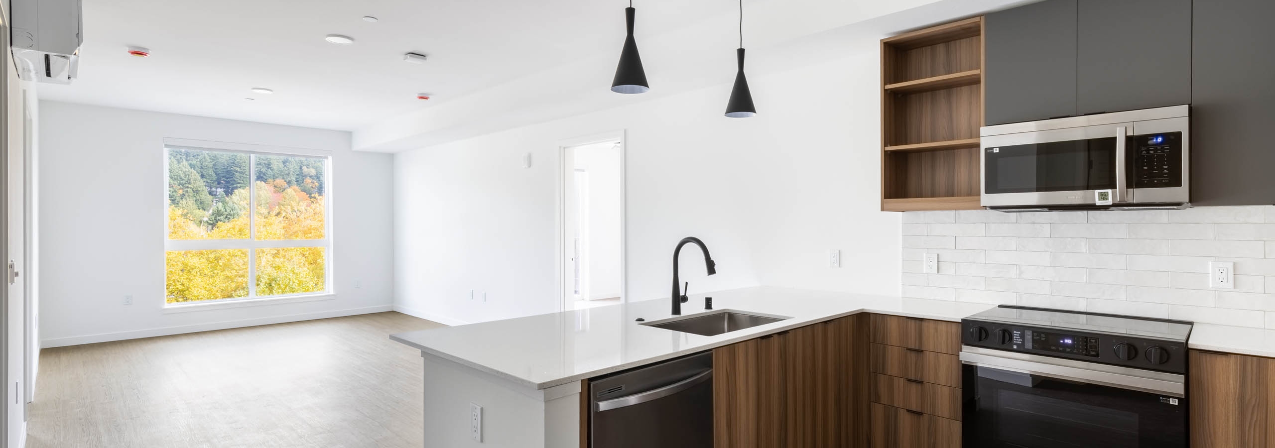 Wide view of AMLI Redmond Way apartment kitchen and living area with pendant lights, wood floors, and large window.