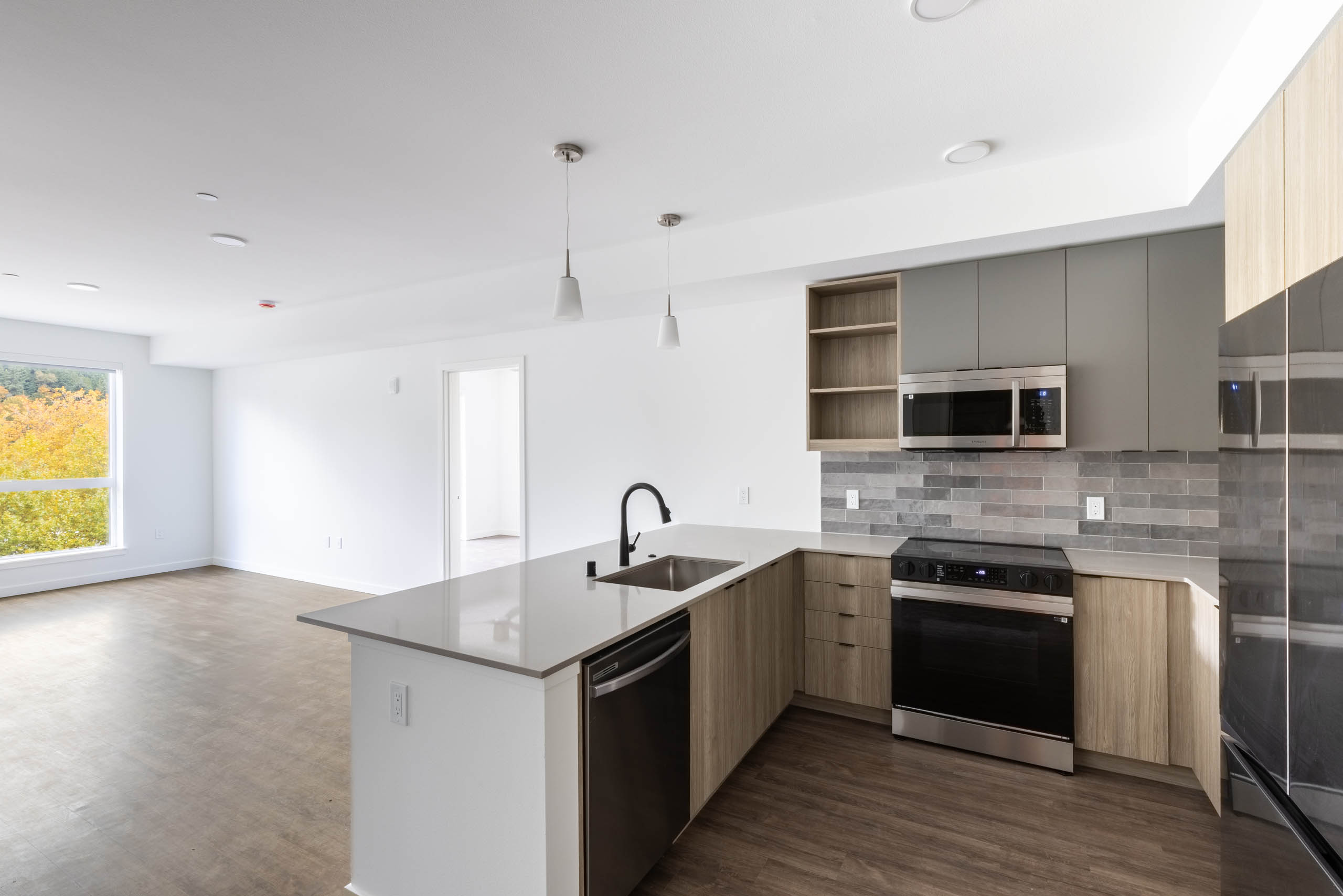 Open view of AMLI Redmond Way apartment kitchen and living space with white pendant lighting, wood floors, and natural window light.