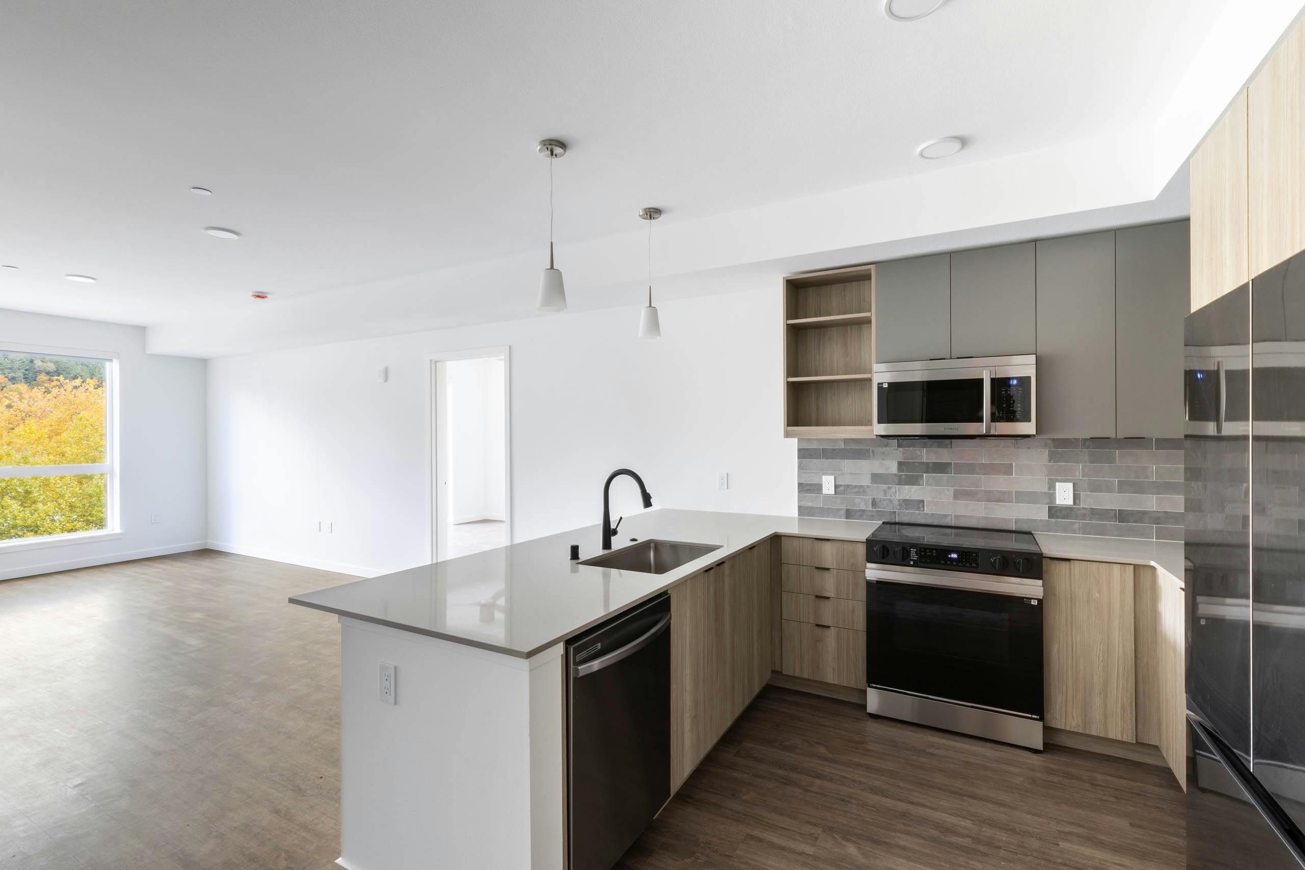 Open view of AMLI Redmond Way apartment kitchen and living space with white pendant lighting, wood floors, and natural window light.