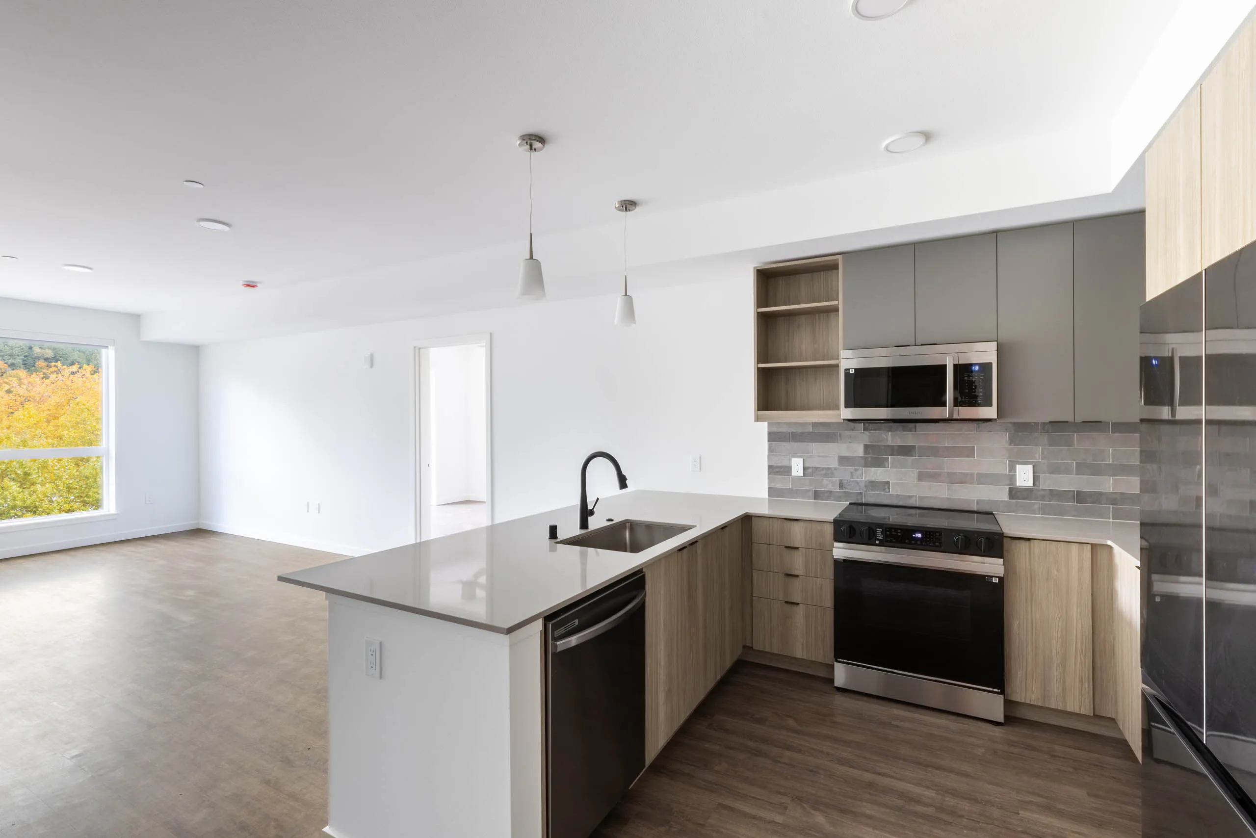 Open view of AMLI Redmond Way apartment kitchen and living space with white pendant lighting, wood floors, and natural window light.