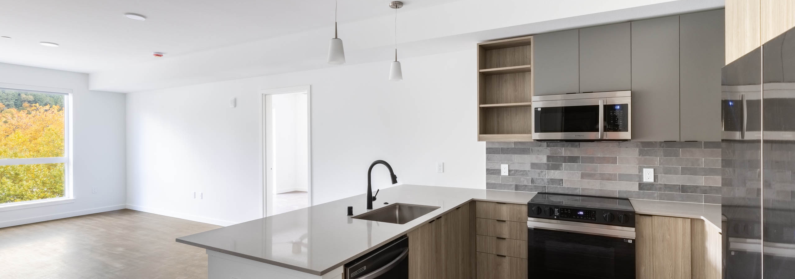 Open view of AMLI Redmond Way apartment kitchen and living space with white pendant lighting, wood floors, and natural window light.