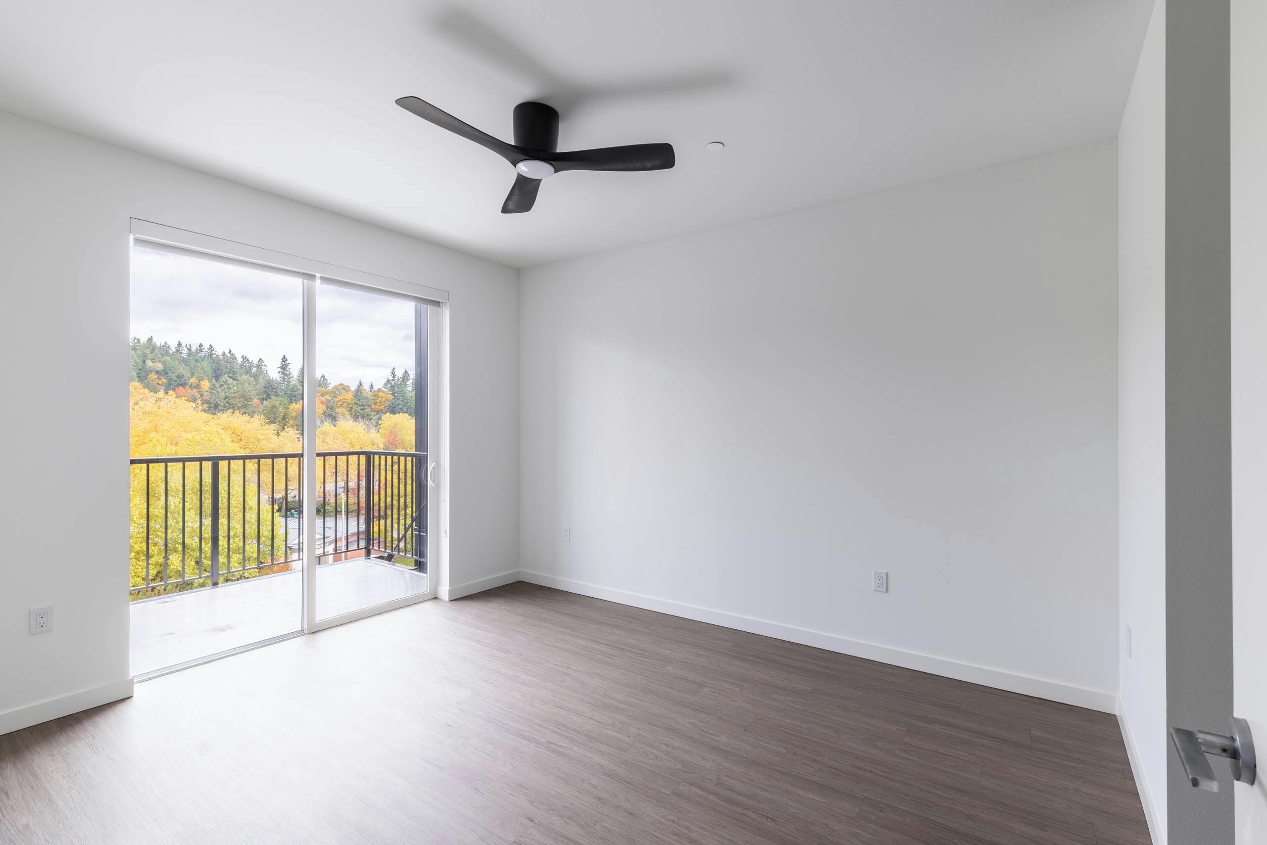 Interior view of AMLI Redmond Way apartment bedroom with ceiling fan, sliding glass door, and private balcony access with forest view.