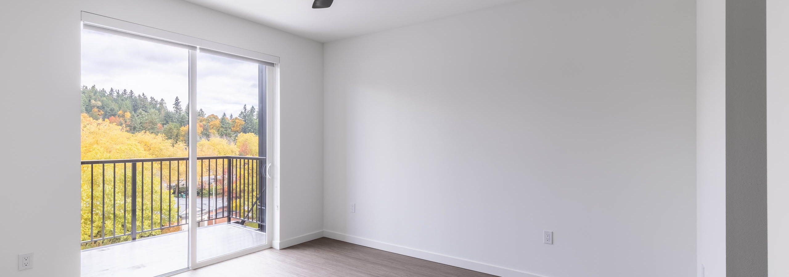 Interior view of AMLI Redmond Way apartment bedroom with ceiling fan, sliding glass door, and private balcony access with forest view.