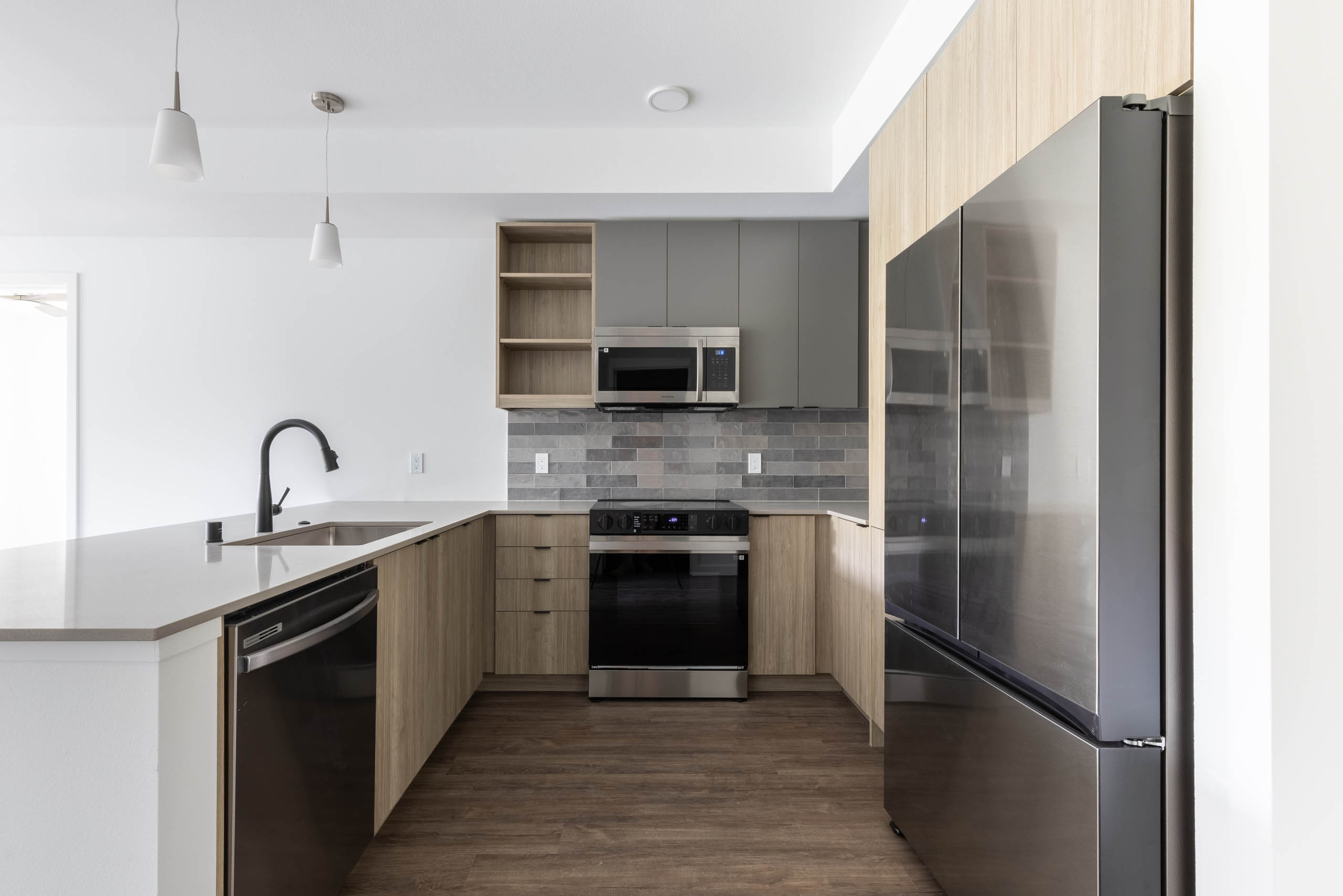Interior view of AMLI Redmond Way apartment kitchen with gray tile backsplash, quartz counters, light wood cabinets and pendant lights.