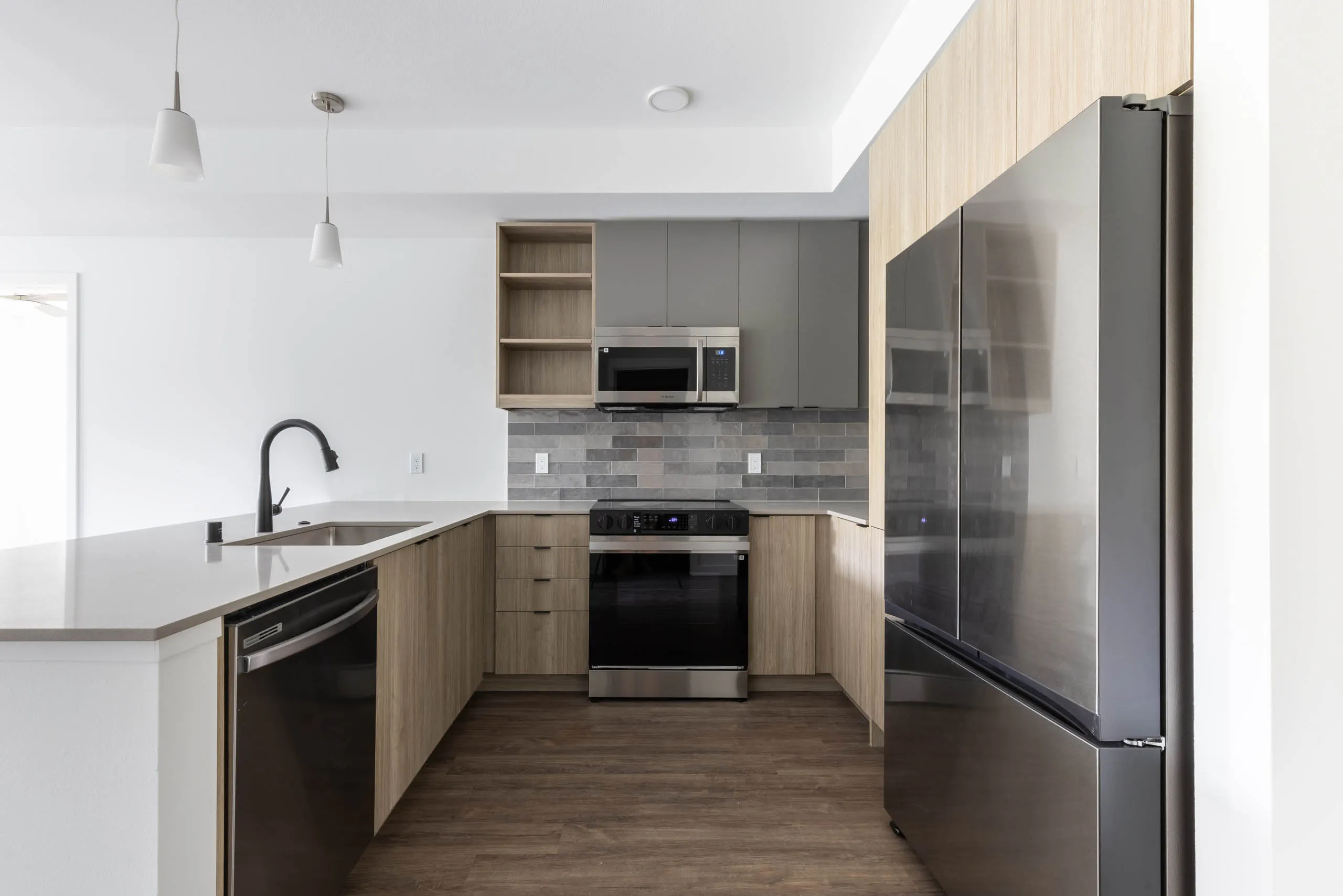 Interior view of AMLI Redmond Way apartment kitchen with gray tile backsplash, quartz counters, light wood cabinets and pendant lights.