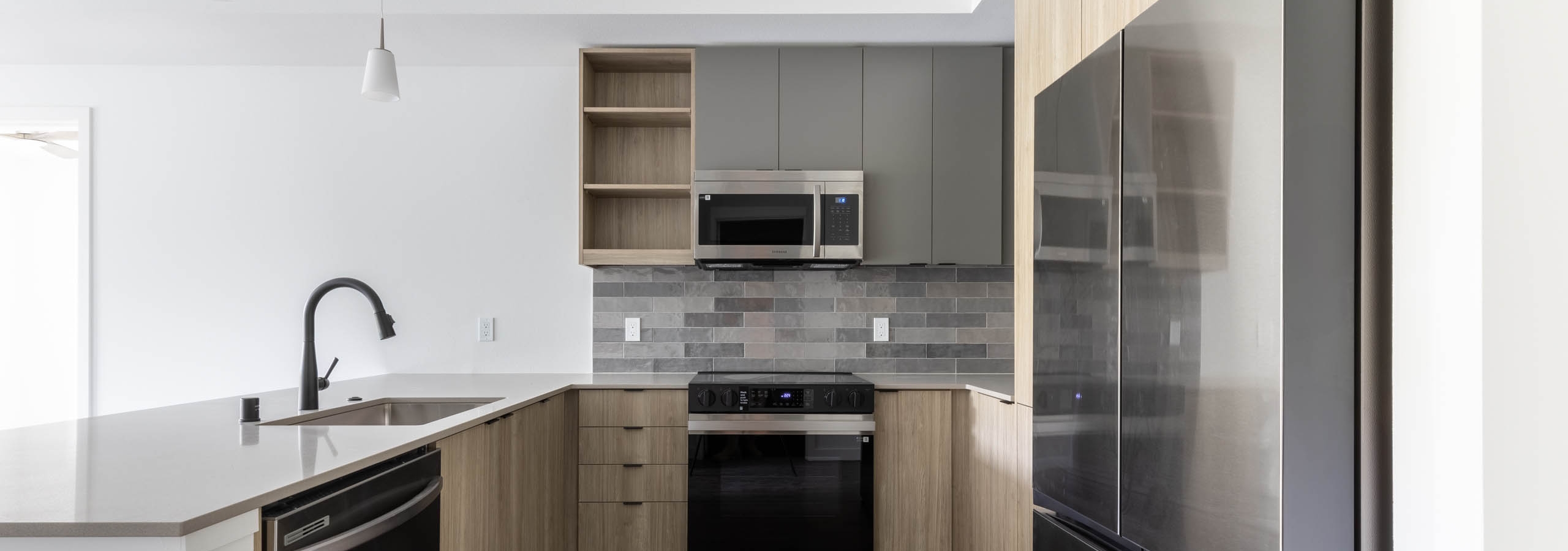 Interior view of AMLI Redmond Way apartment kitchen with gray tile backsplash, quartz counters, light wood cabinets and pendant lights.