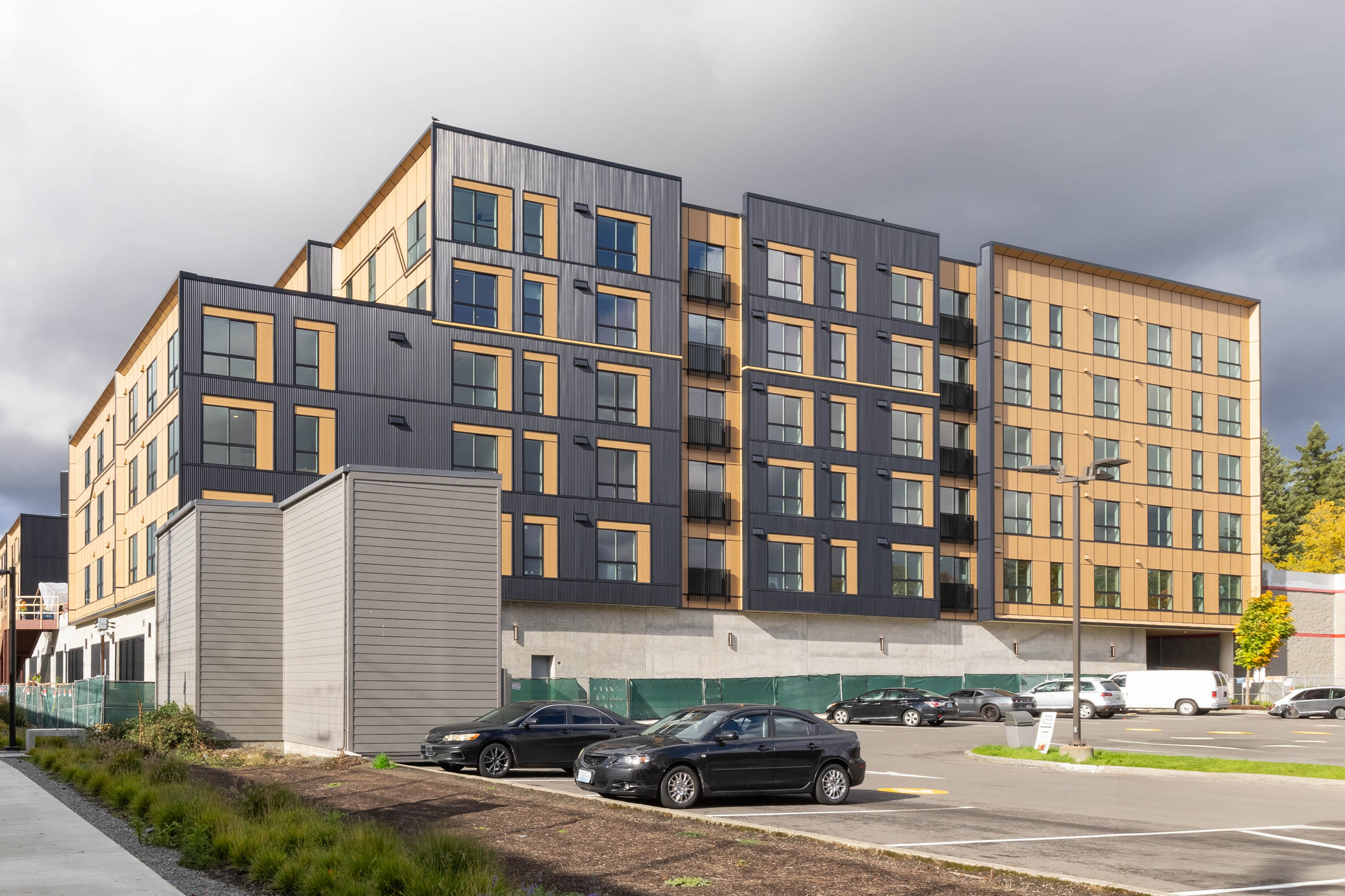 Daytime exterior of AMLI Redmond Way apartment facade with black and tan panels, parked cars, and nearby pedestrian path.
