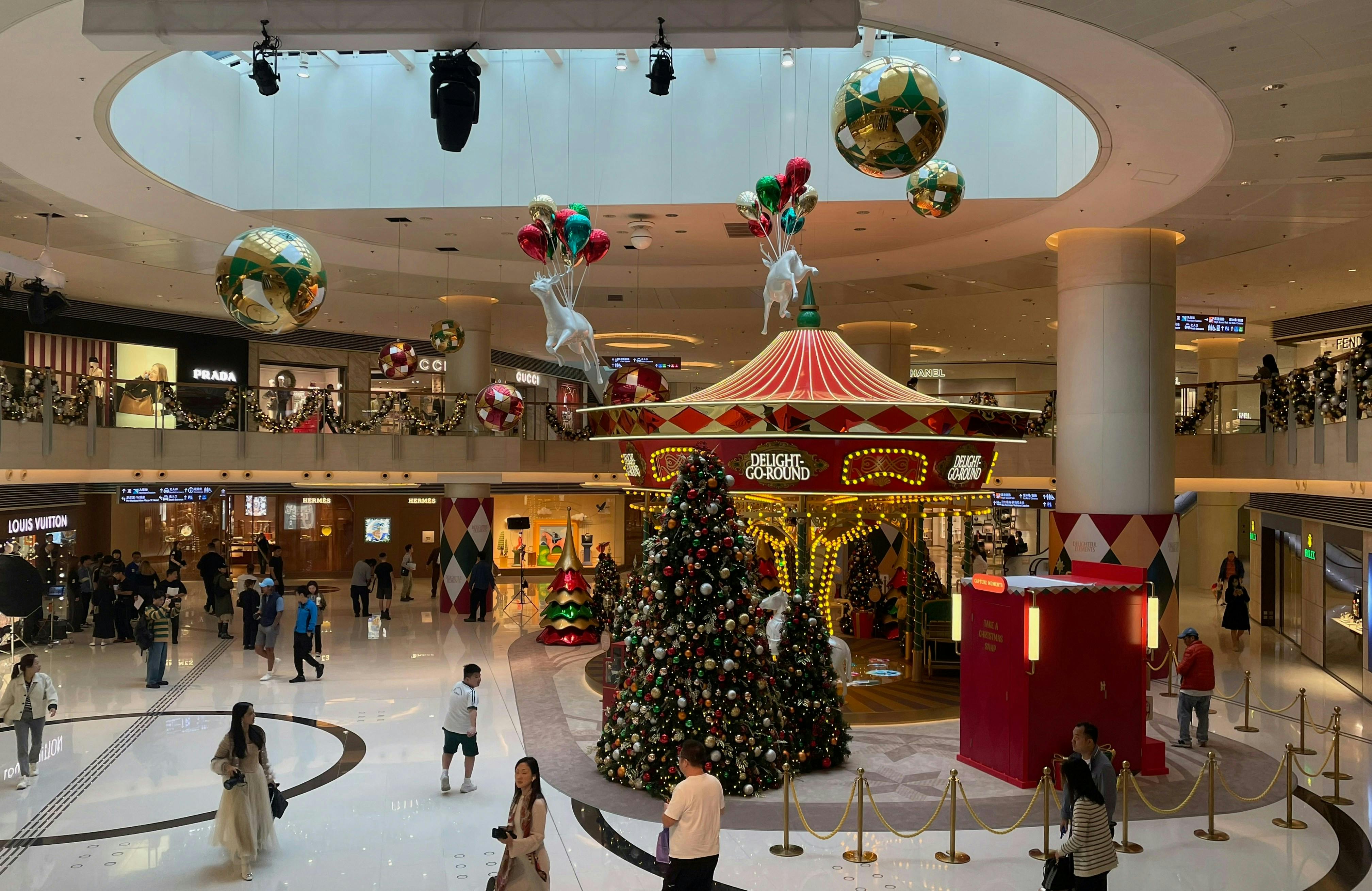 A festive, brightly lit shopping mall atrium featuring a large, traditional Christmas tree and a brightly colored seasonal carousel in the center. Large, decorative ornaments hang from the high ceiling, and upscale store facades like Prada and Louis Vuitton line the multiple levels.