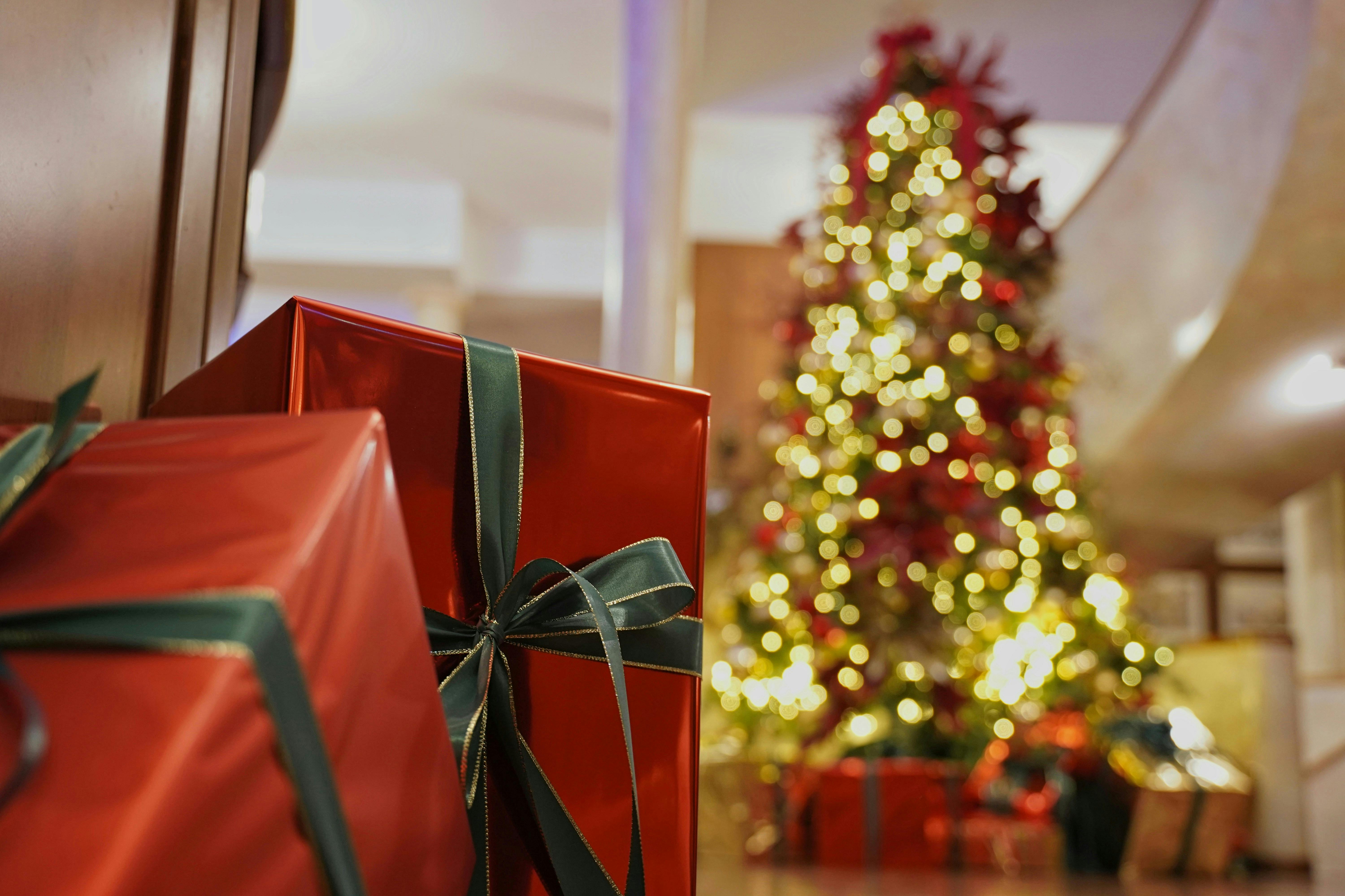 A shallow-focus shot featuring large red Christmas presents wrapped with dark green ribbons in the foreground. In the background, a large, brightly lit, blurred Christmas tree with gold and red lights provides a warm, festive glow.