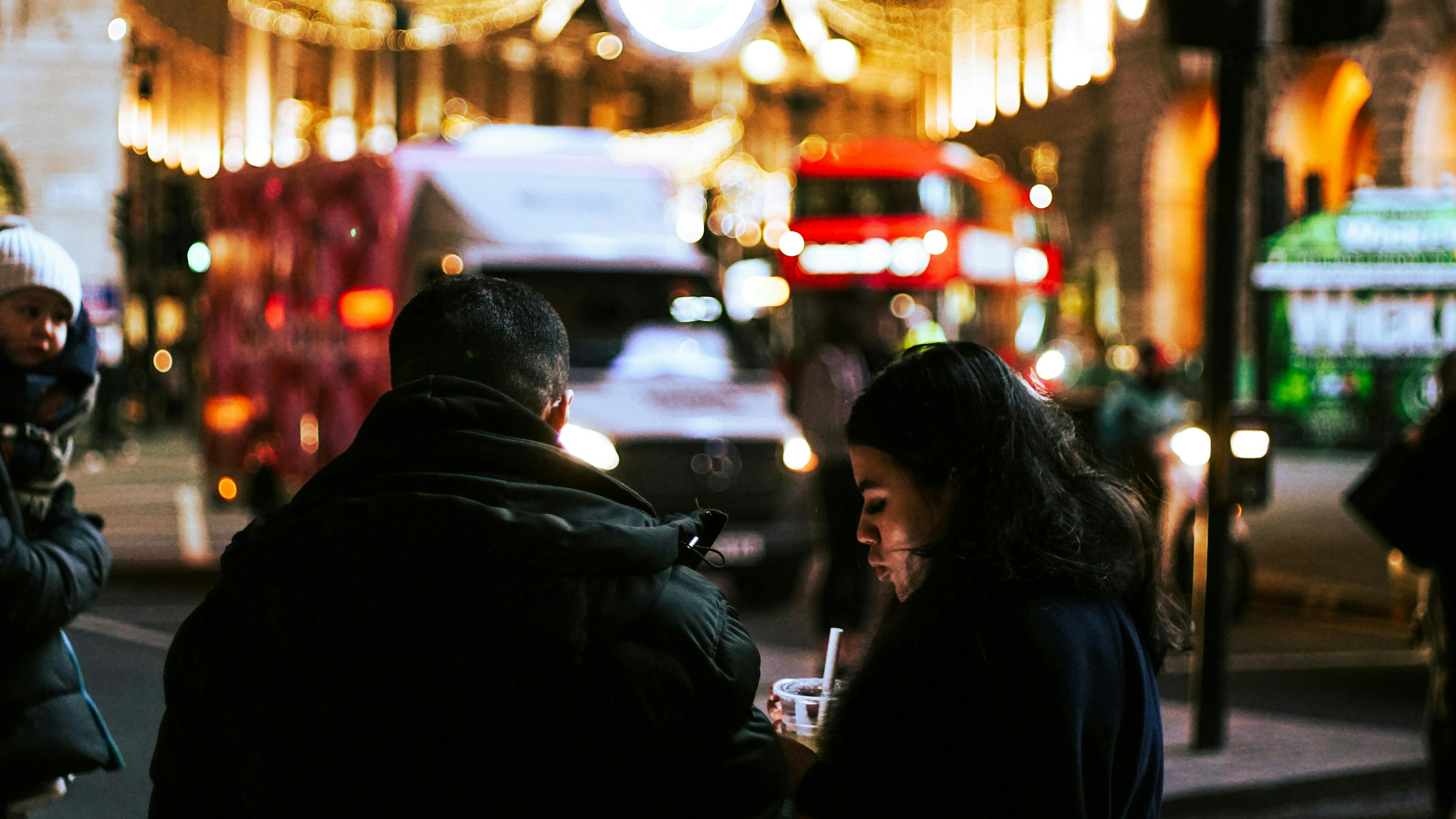 A tightly focused, low-light shot of the backs of two people standing on a city street at night. A woman on the right sips a drink with a straw. The background is a vibrant blur of red buses, cars, and golden holiday lights strung across the street.