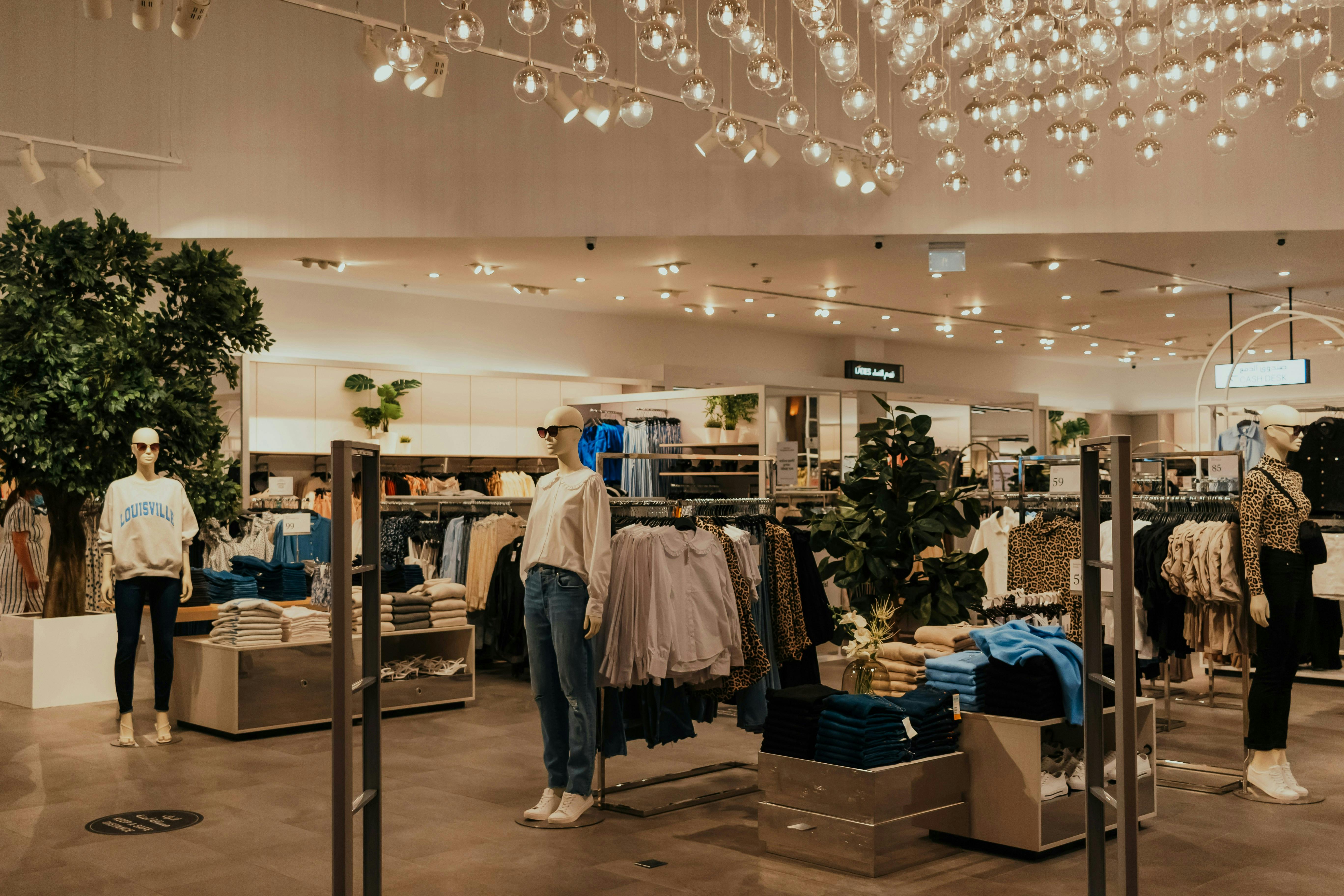 The warm and inviting interior of a clothing store. Mannequins wearing casual attire stand among displays of folded and hanging clothes. Large indoor trees and numerous small, suspended globe light fixtures contribute to a cozy and contemporary atmosphere.