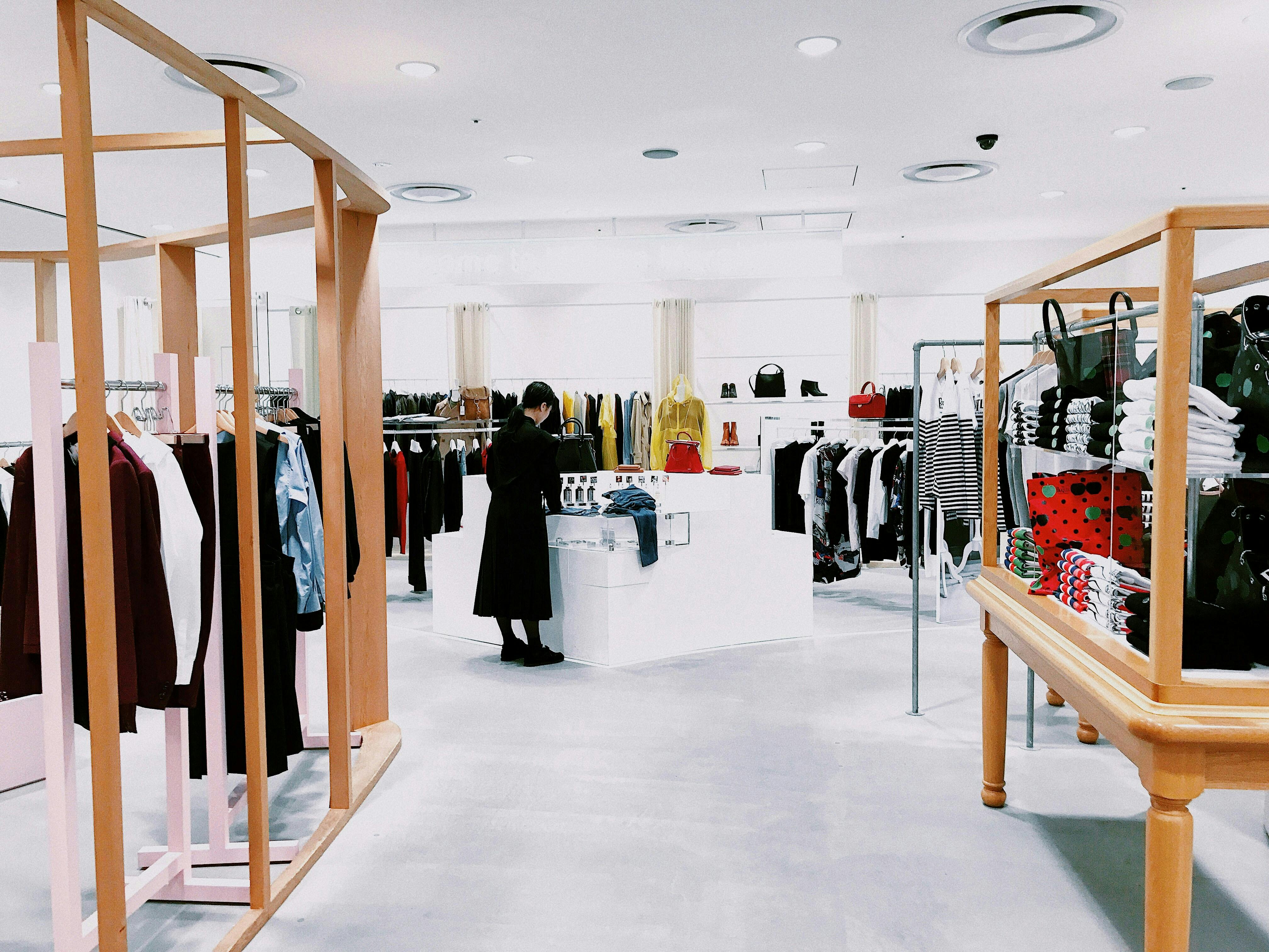 A brightly lit, minimalist clothing store with white walls and flooring. A person in a long black coat is examining merchandise on a white central display table. Clothing racks framed by blonde wooden structures are on the sides, featuring a variety of colorful apparel.