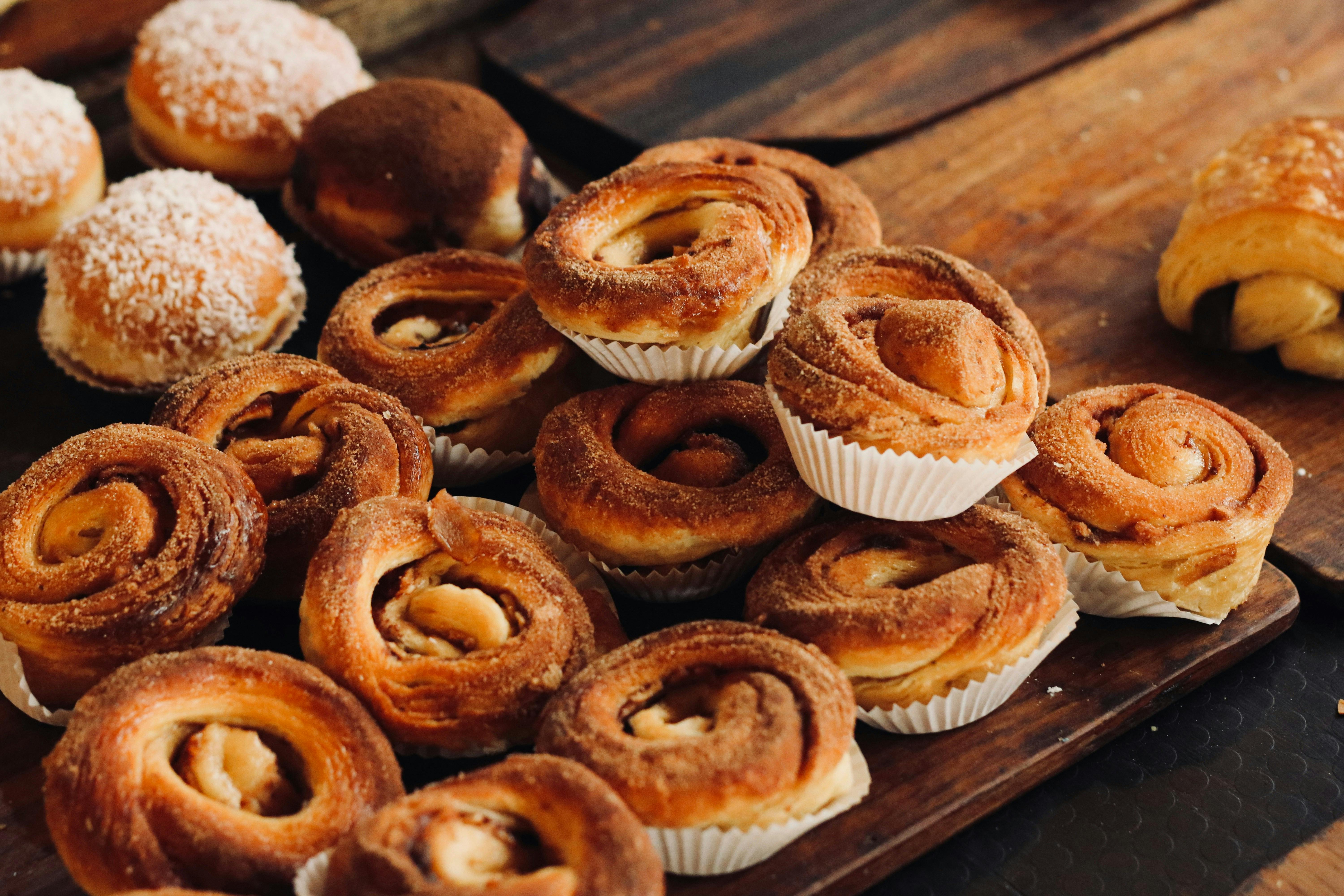 An arrangement of various pastries on a dark wooden board. The majority are small, spiral cinnamon buns in white paper liners, sprinkled with cinnamon. In the background, there are round doughnuts, some covered in powdered sugar and others with cocoa powder.