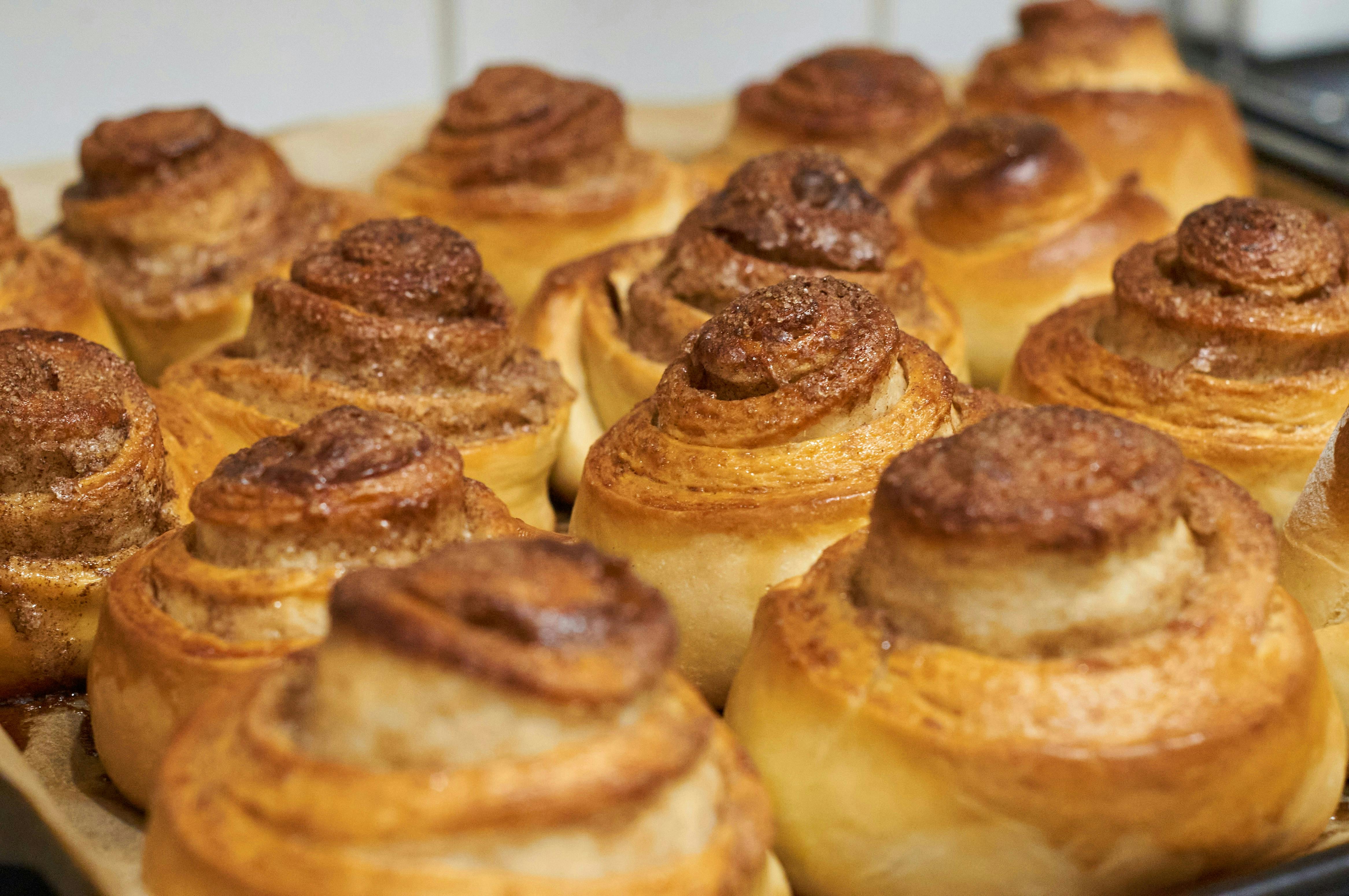 A close-up shot of a baking sheet filled with freshly baked, golden-brown cinnamon rolls right out of the oven. The tops are glossy and slightly caramelized with a visible sprinkle of cinnamon sugar.