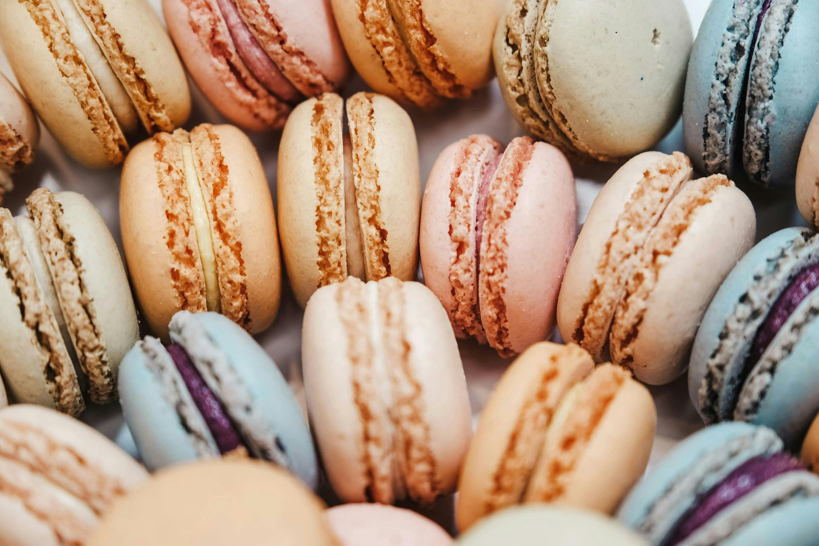 A macro, overhead view of a collection of brightly colored French macarons. The flavors include shades of pale pink, light blue, cream, and tan, showcasing the delicate, cracked shells and creamy filling.
