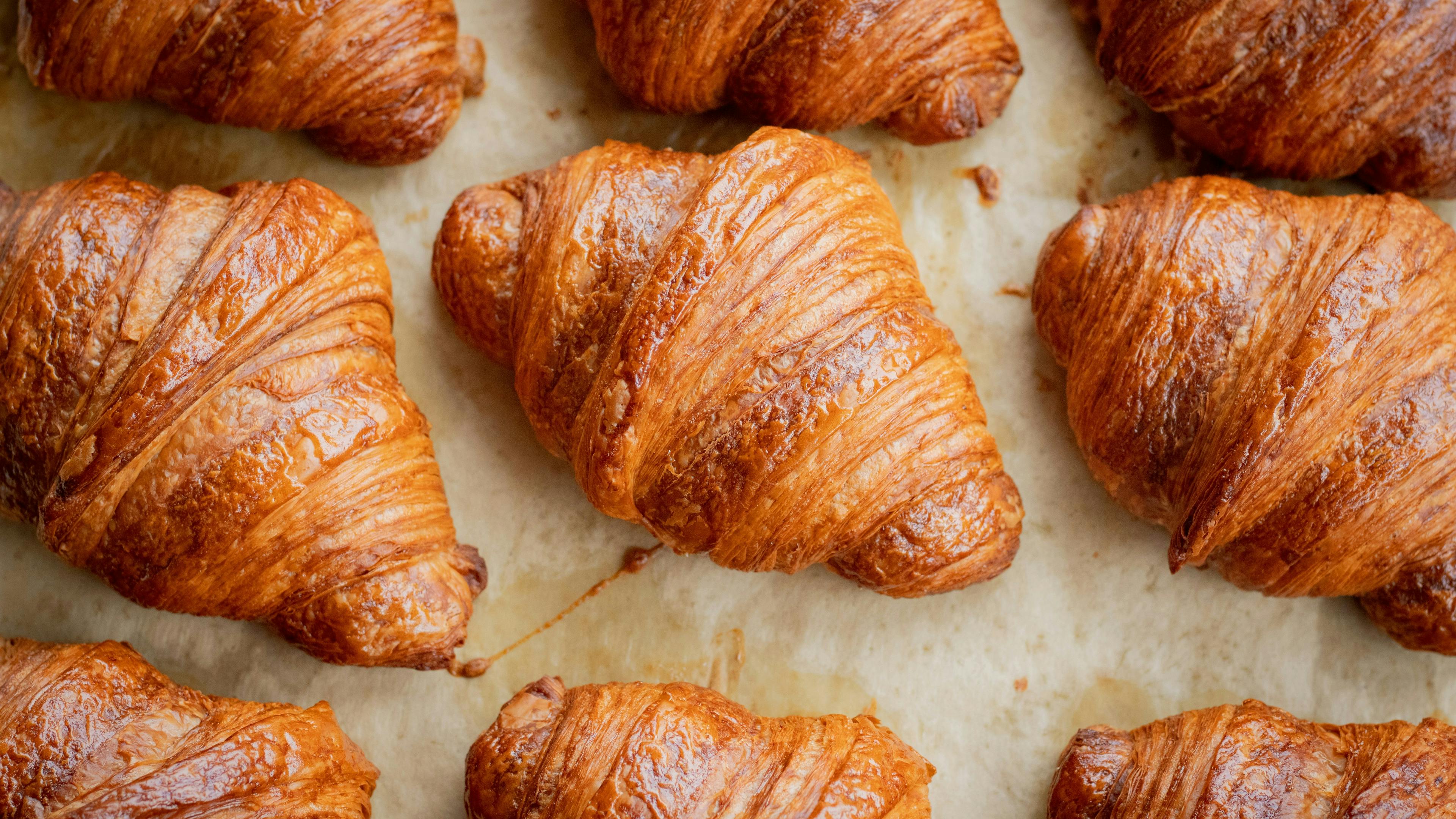 A top-down, close-up view of several freshly baked croissants arranged on a piece of parchment paper. The pastries have a rich, golden-brown crust with visible flaky layers.