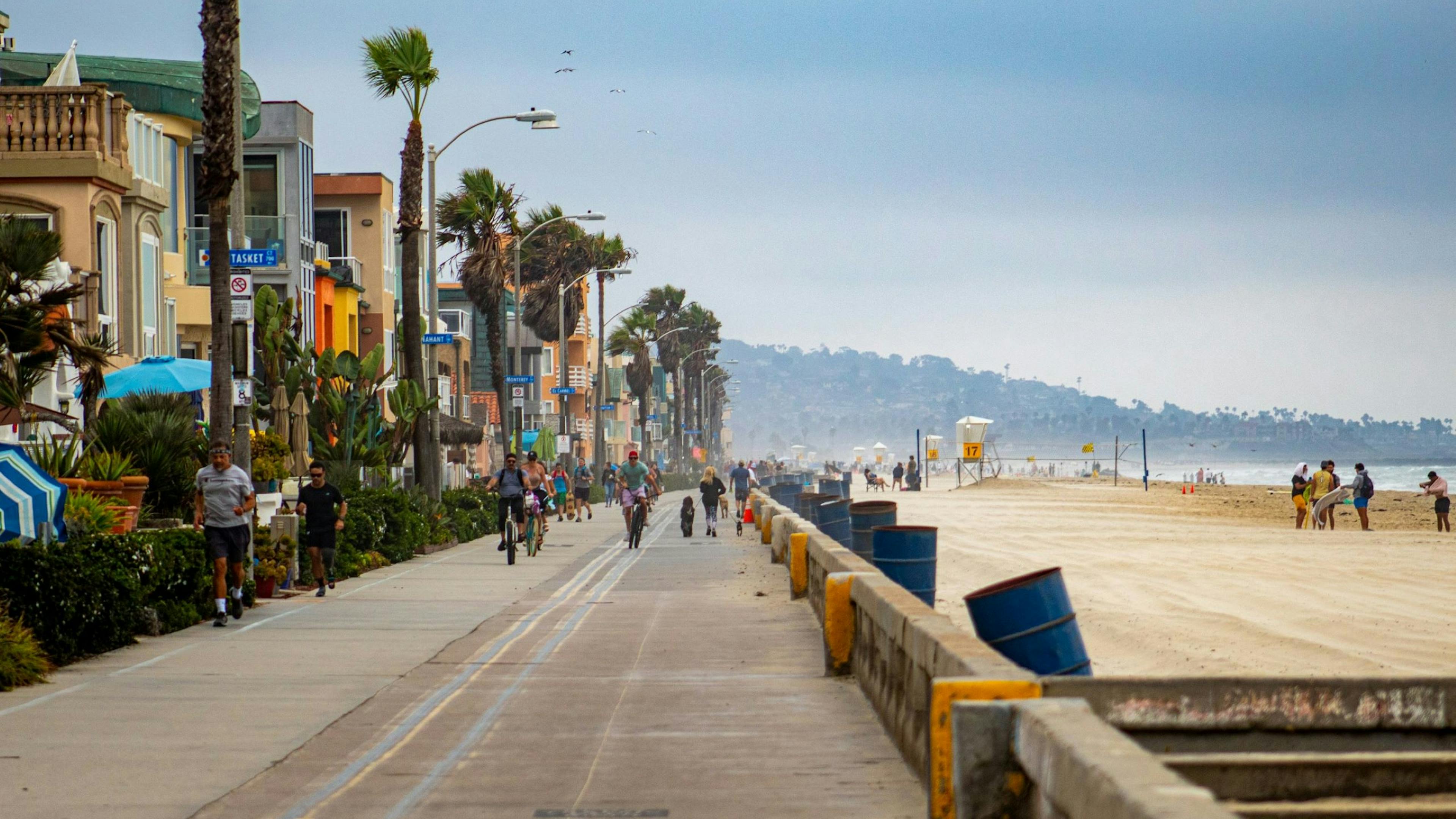 People walking, jogging, and biking on a beachside promenade with palm trees and beachfront homes on a cloudy day.