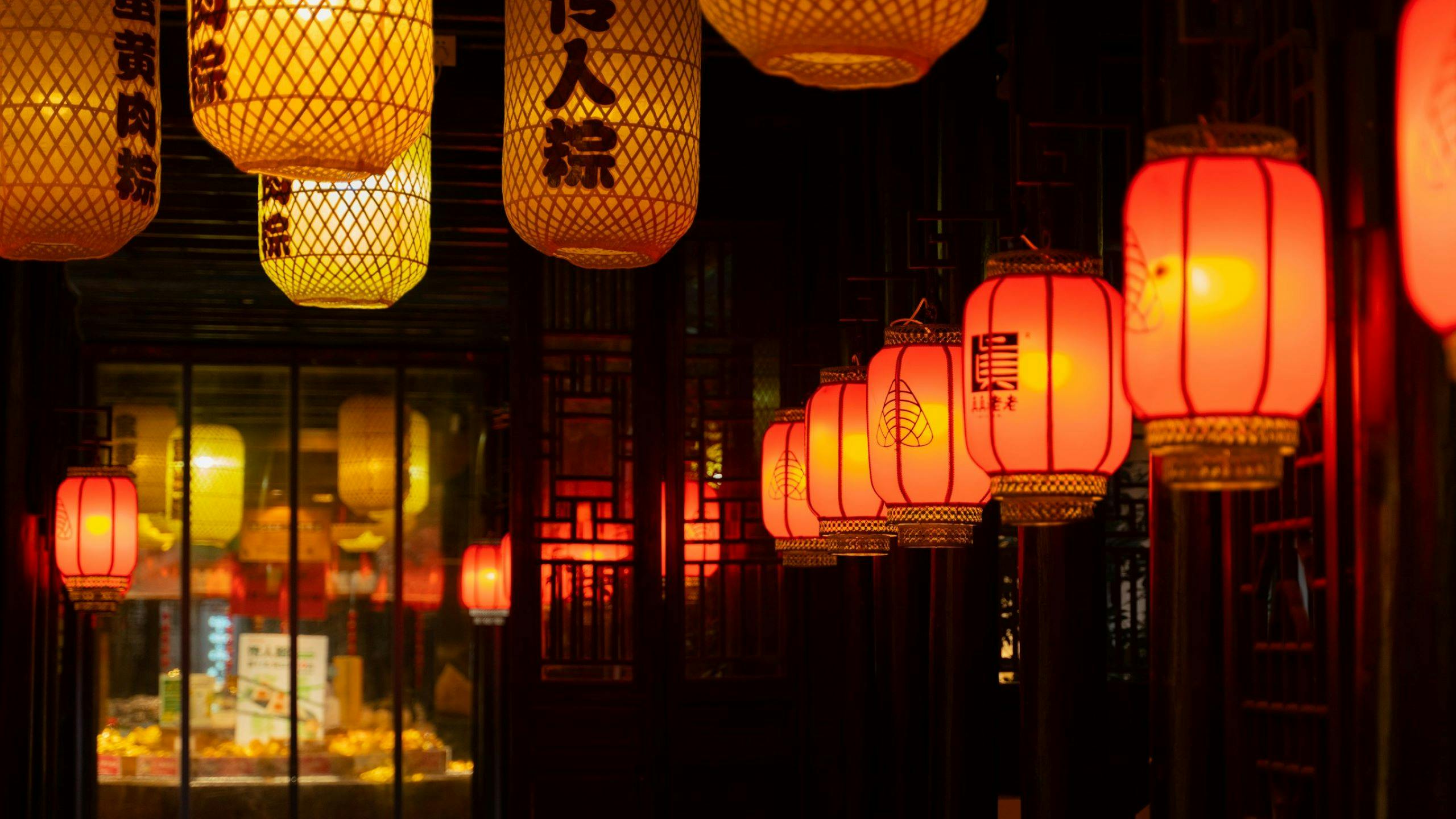 Lanterns, both woven yellow and round red, illuminate a dark interior space.
