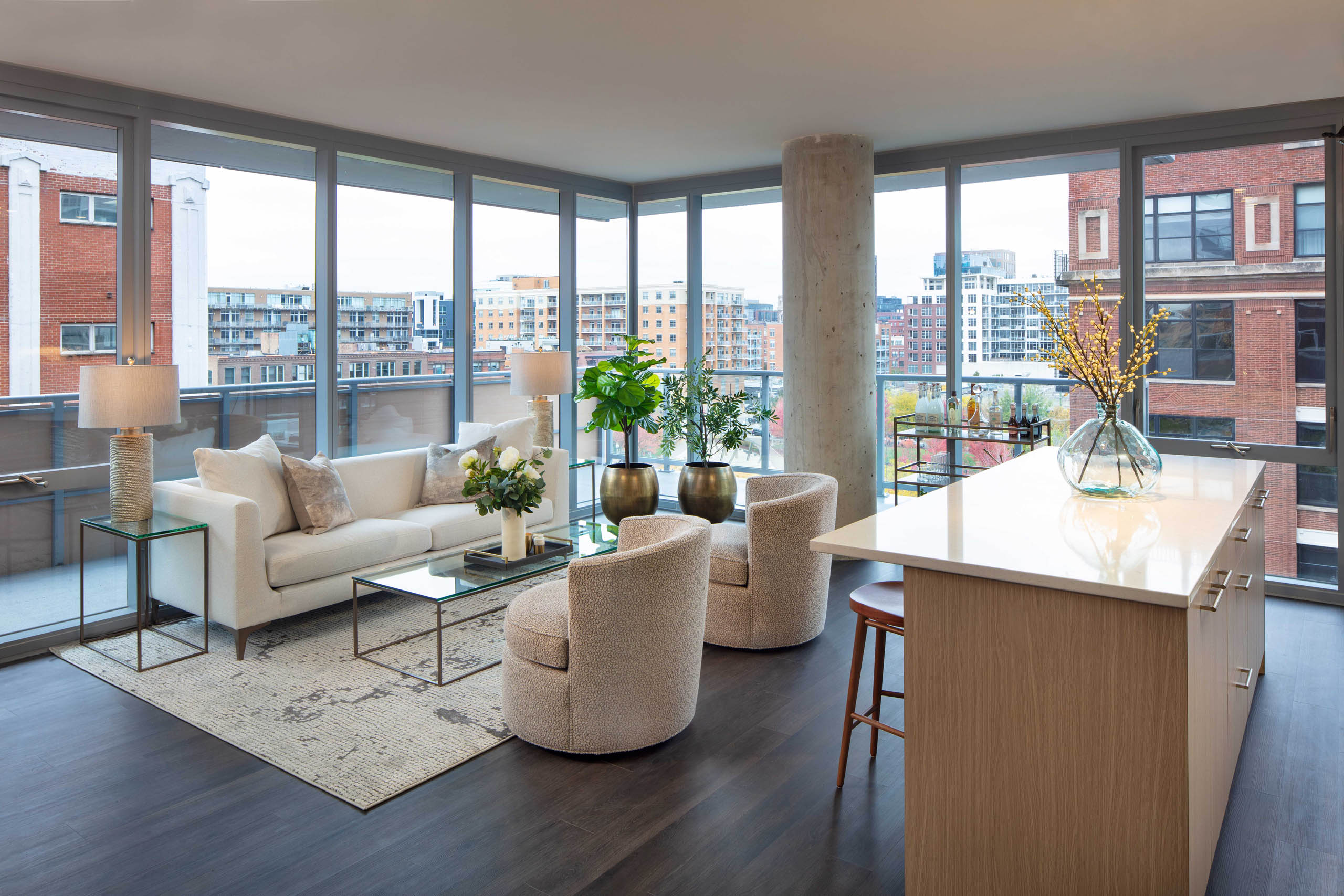 Living room with a white sofa, two textured chairs, a kitchen island, dark flooring, and floor-to-ceiling windows with a city view at AMLI West Loop