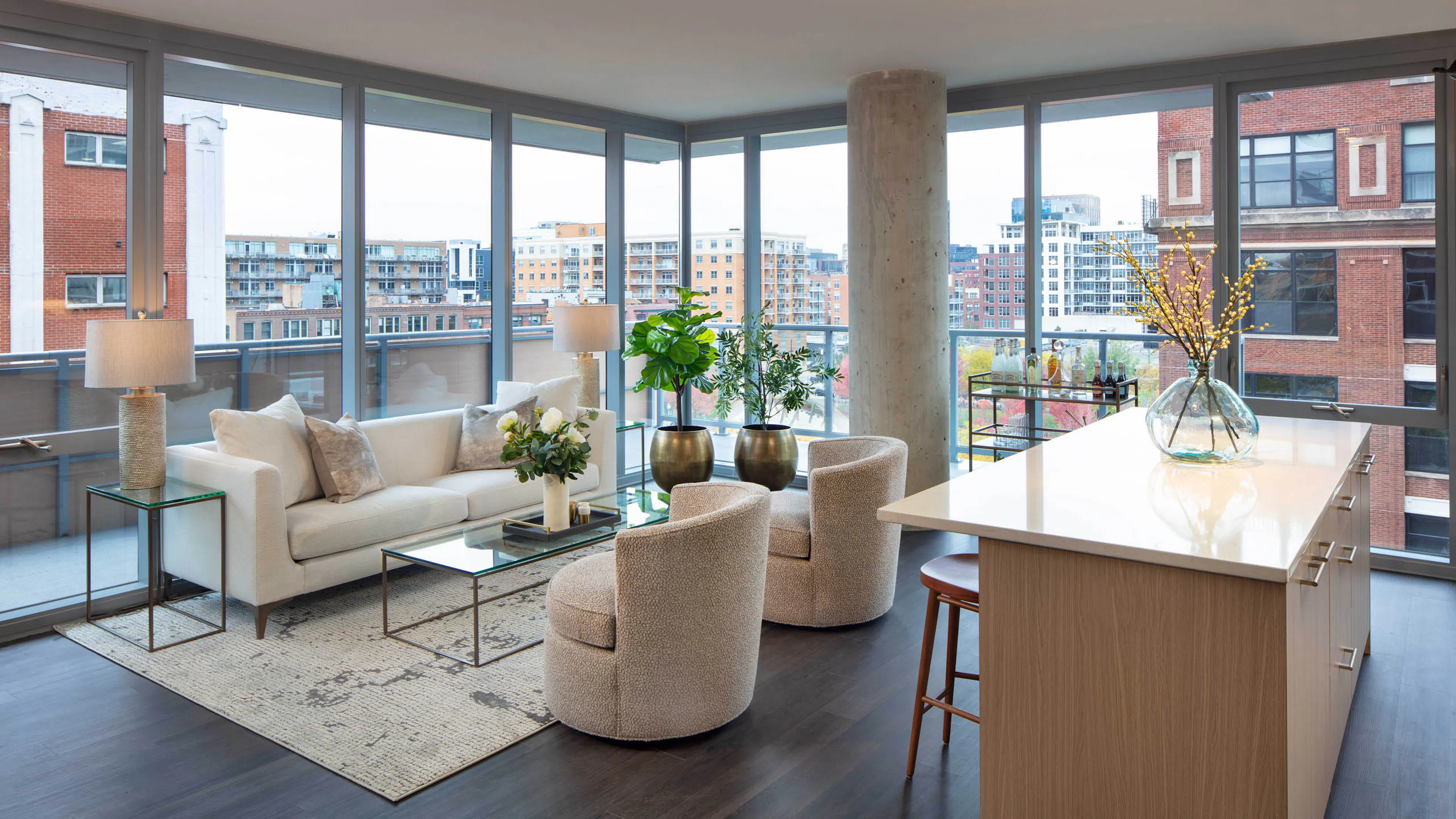Living room with a white sofa, two textured chairs, a kitchen island, dark flooring, and floor-to-ceiling windows with a city view at AMLI West Loop