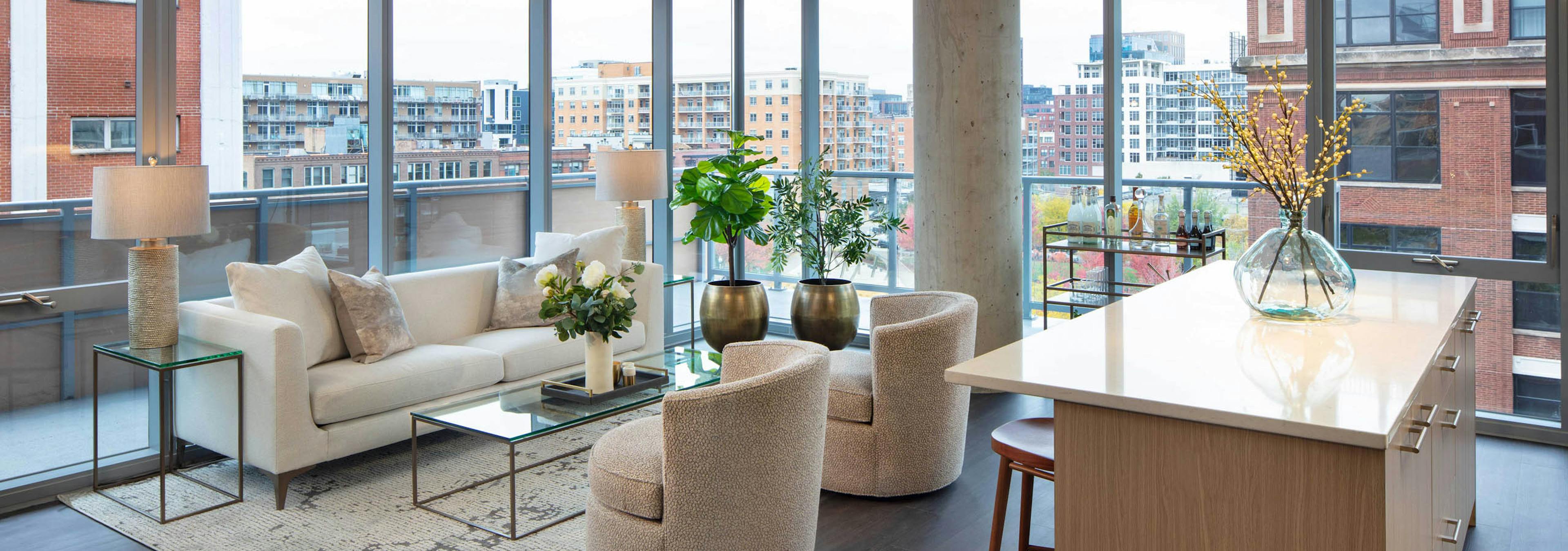 Living room with a white sofa, two textured chairs, a kitchen island, dark flooring, and floor-to-ceiling windows with a city view at AMLI West Loop